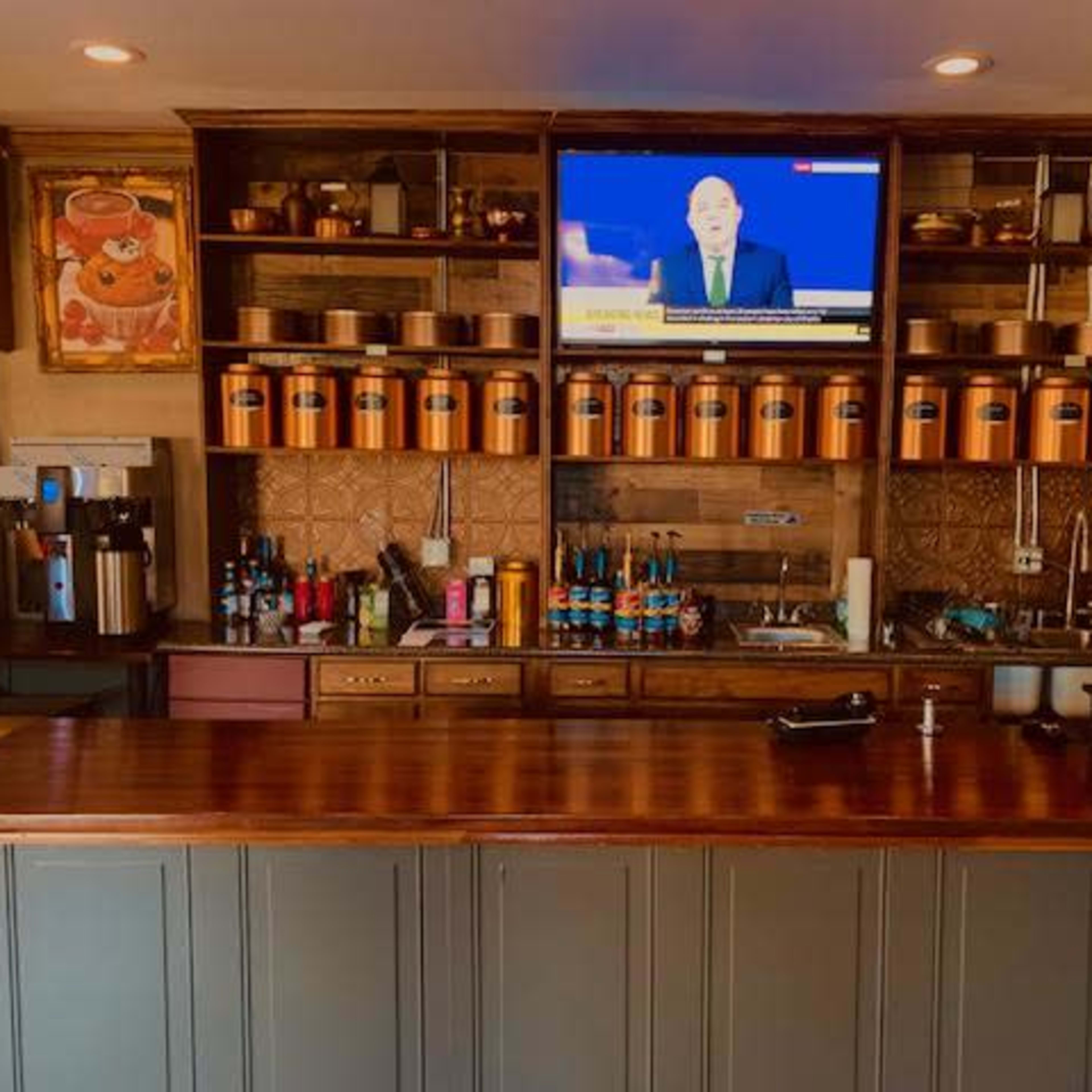 A coffee bar with a wooden countertop, shelves filled with beverage supplies, and a television displaying news in the background.