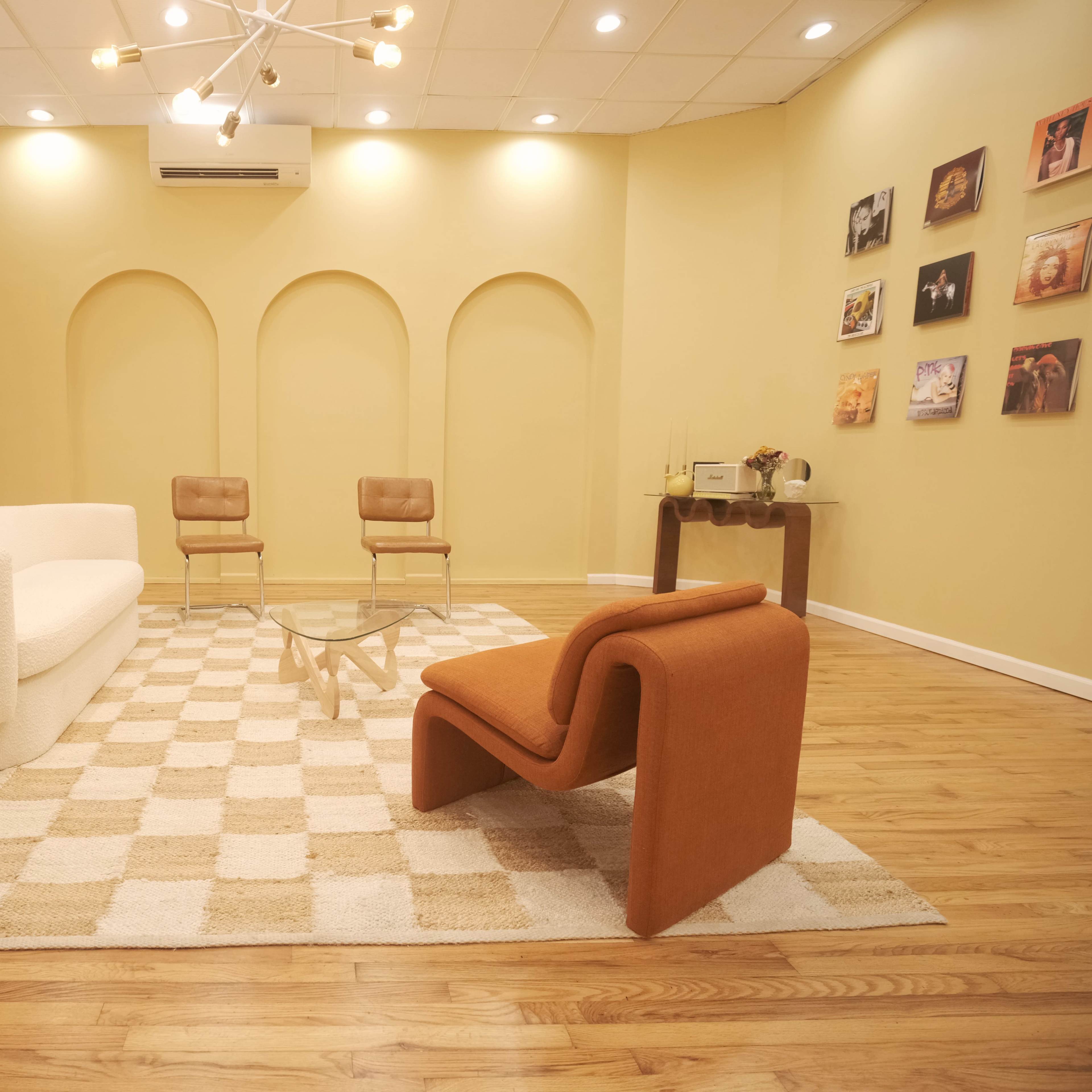 The scene features a modern interior with a curved white sofa, an orange chair, two tan chairs, a glass coffee table, and a checkered rug on a wooden floor, set against yellow walls with art displayed.