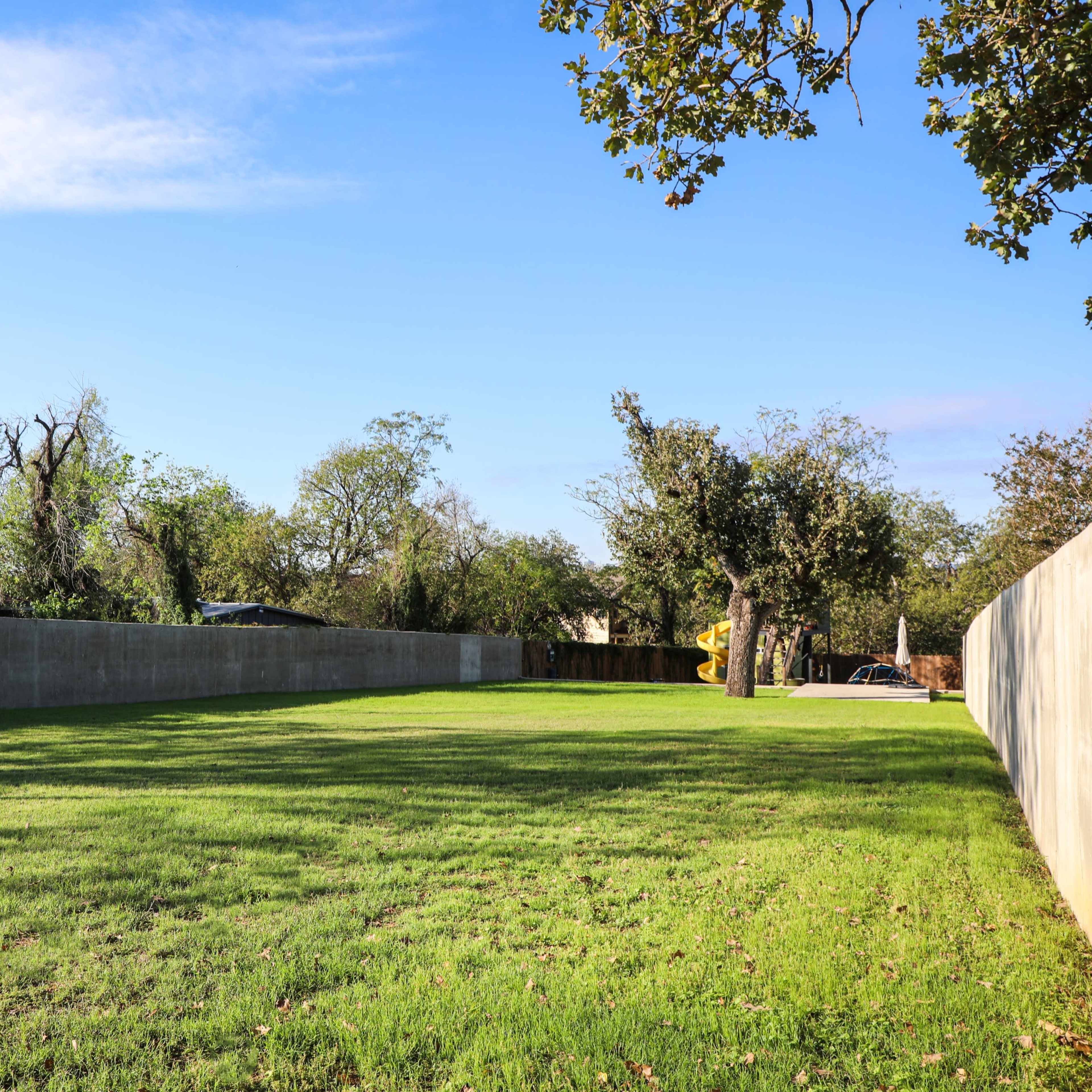 A wide, open grassy area bordered by a concrete wall and trees under a clear blue sky.