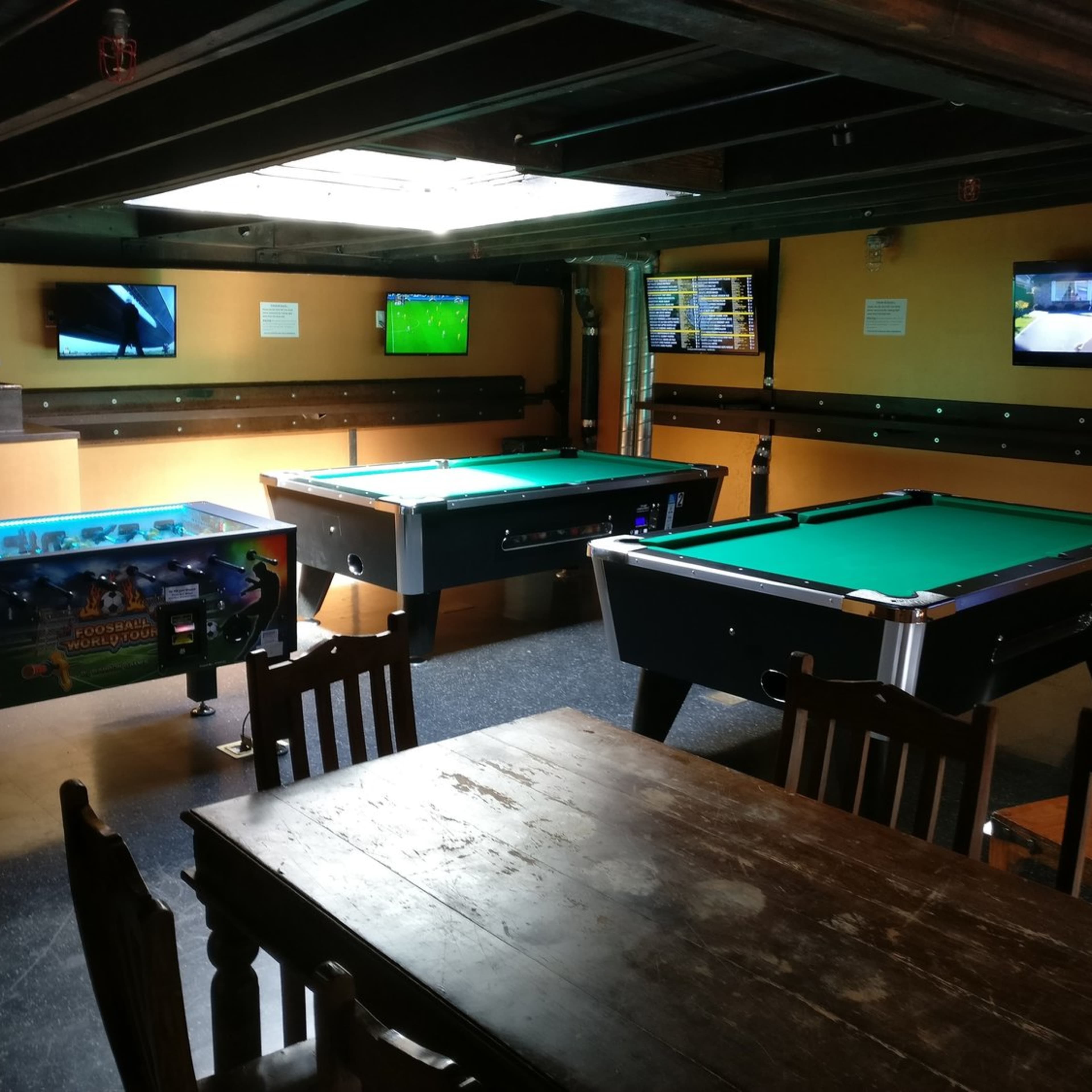 A game room with two pool tables, a pinball machine, and multiple television screens mounted on the walls.