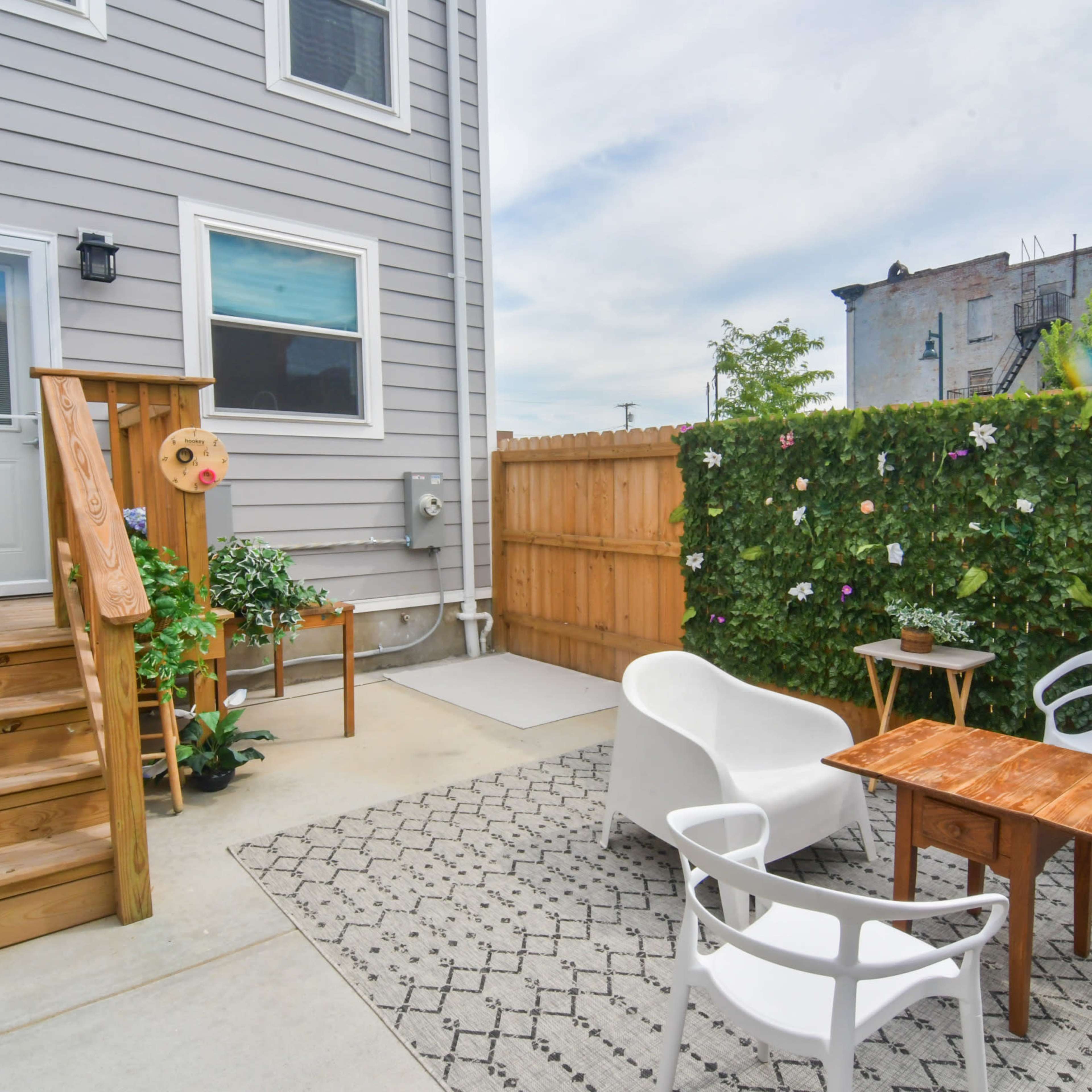 A small outdoor patio area with white chairs and a wooden table, surrounded by a green wall and steps leading to a door.