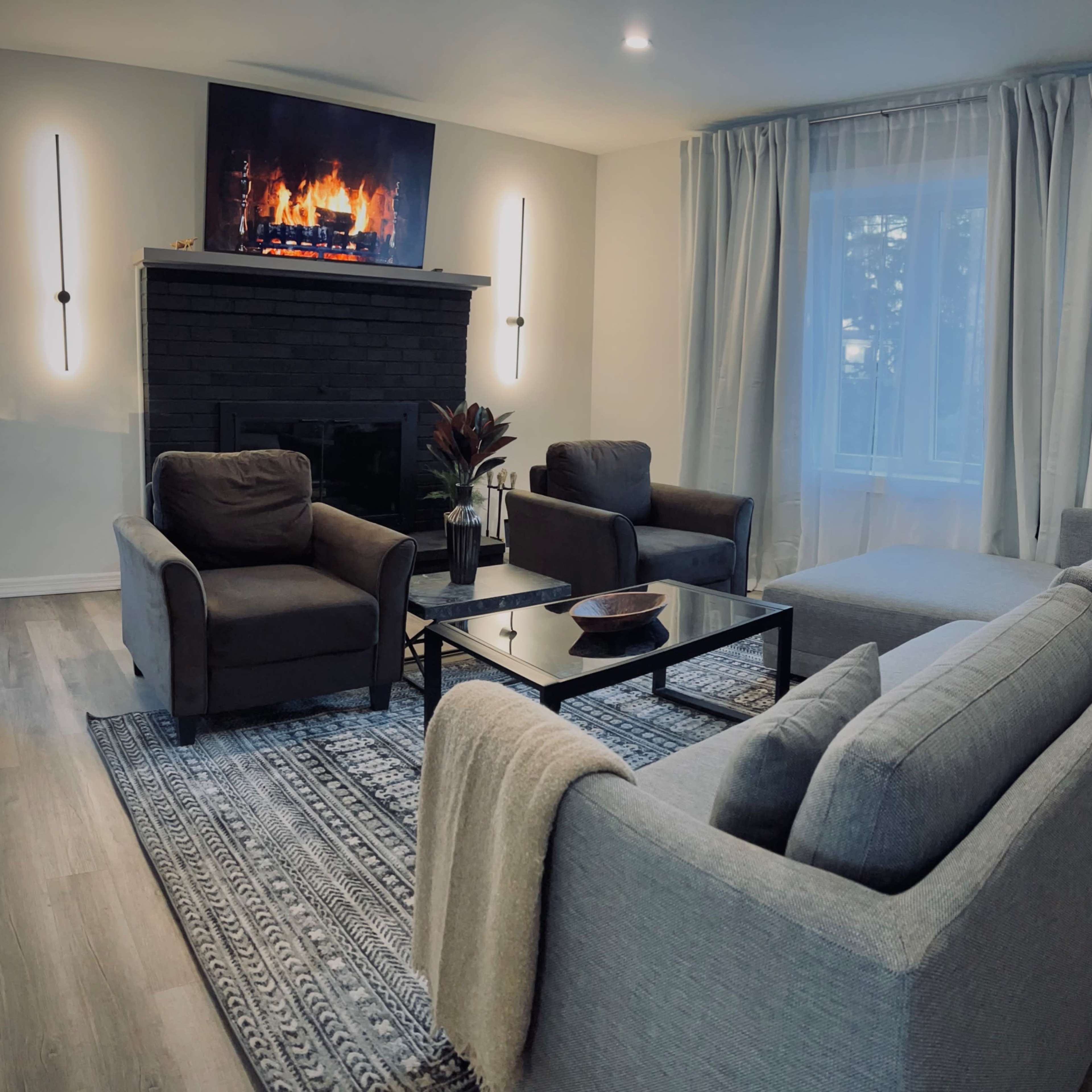 A modern living room with a gray sofa, two armchairs, a coffee table, and a television mounted above a black fireplace.
