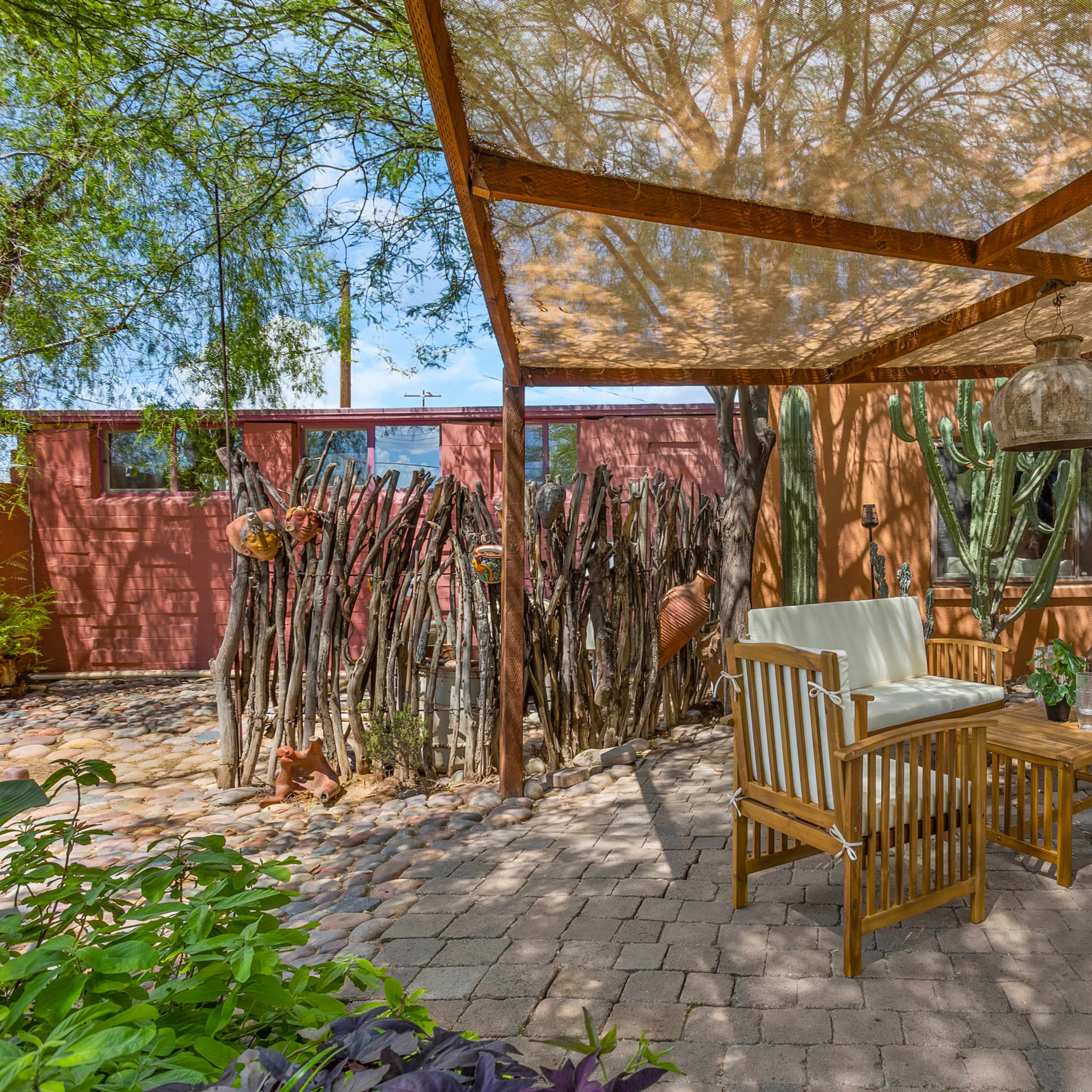 A shaded patio area features wooden chairs and a table, surrounded by a variety of plants and a decorative wall.