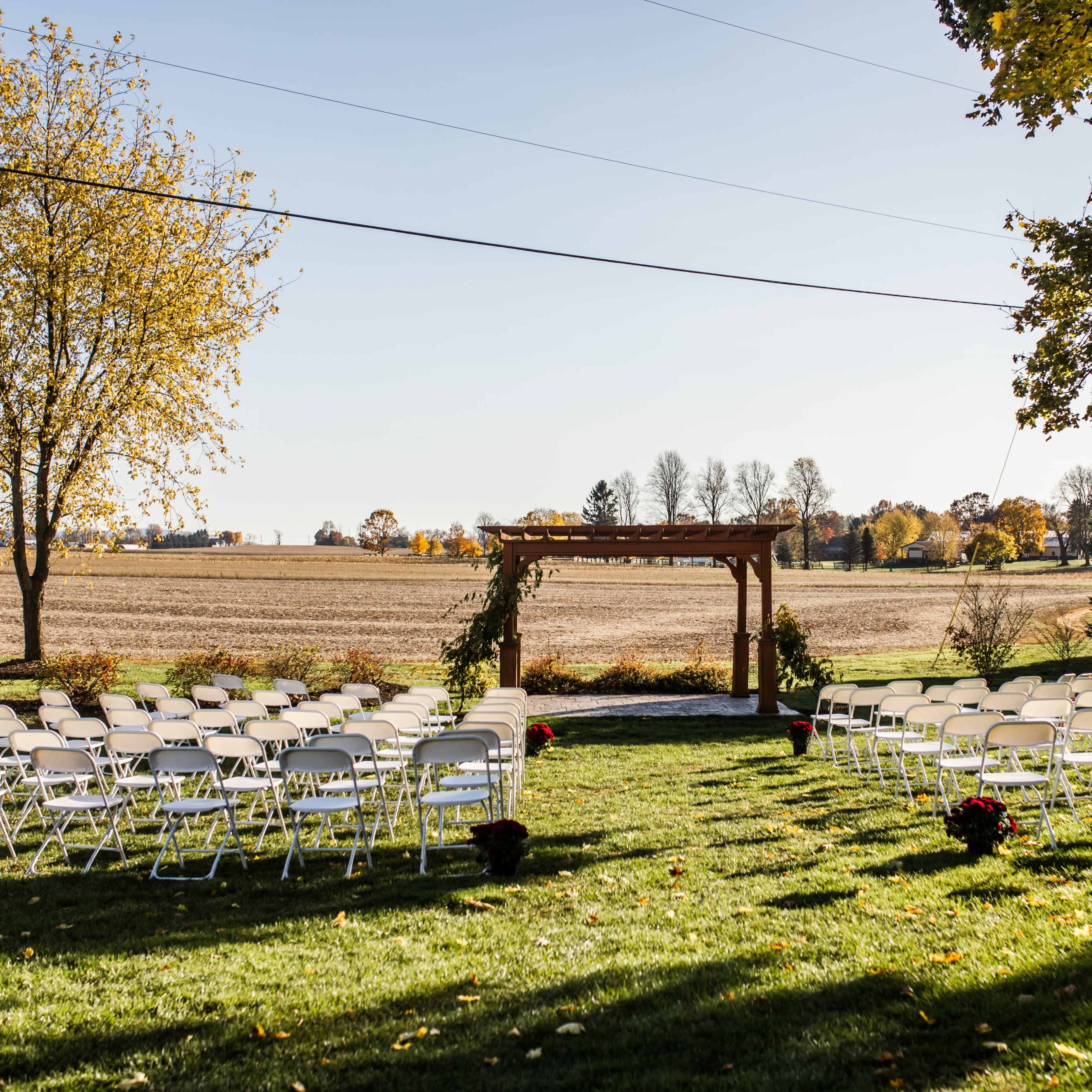 A wedding ceremony setup with rows of white chairs arranged on a grassy area in front of a wooden arbor, overlooking a field and trees in the background.