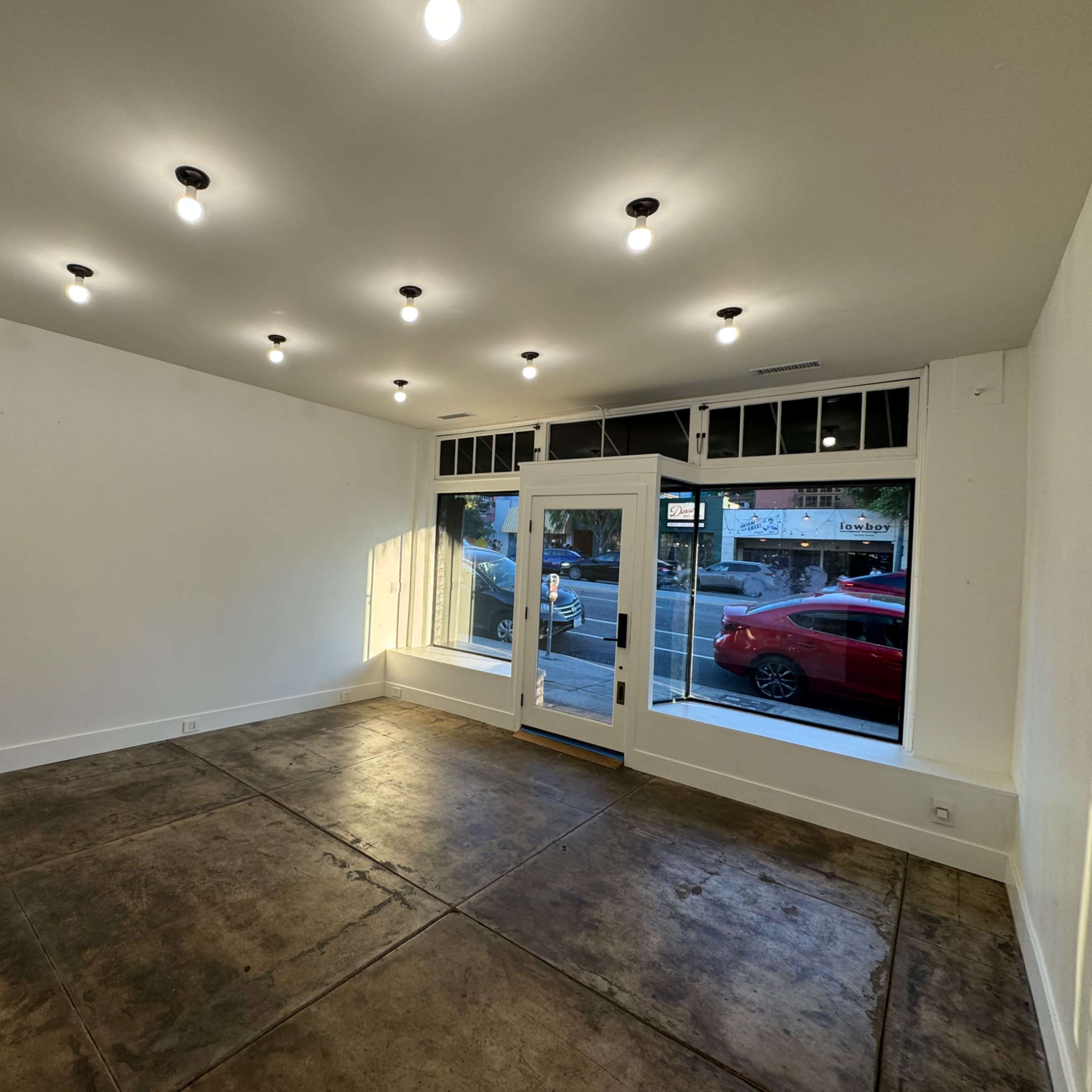 An empty room with a concrete floor and bright lighting, featuring a large front window and a glass door leading outside.