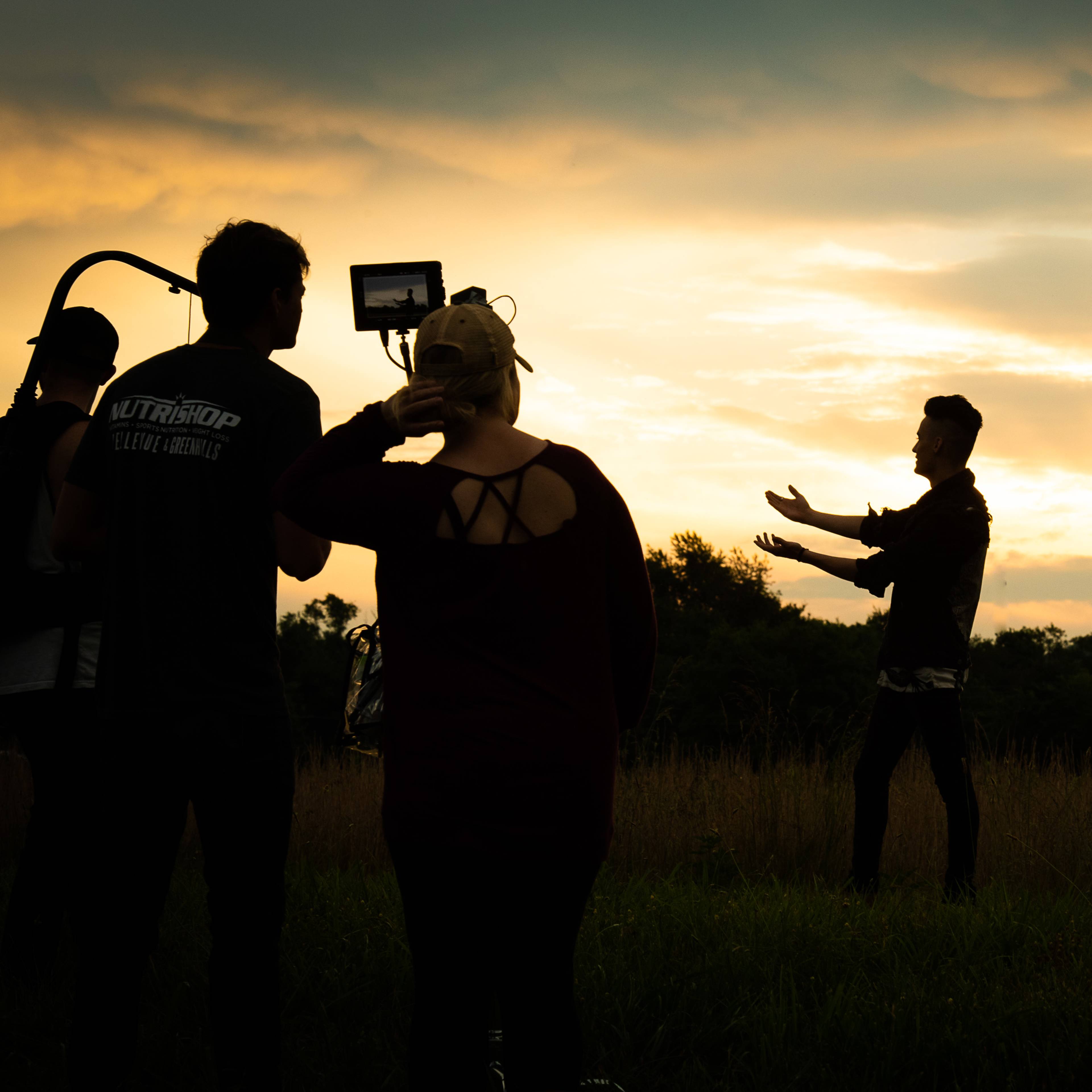 A group of people stands in a field as a person gestures towards the sunset in the background.