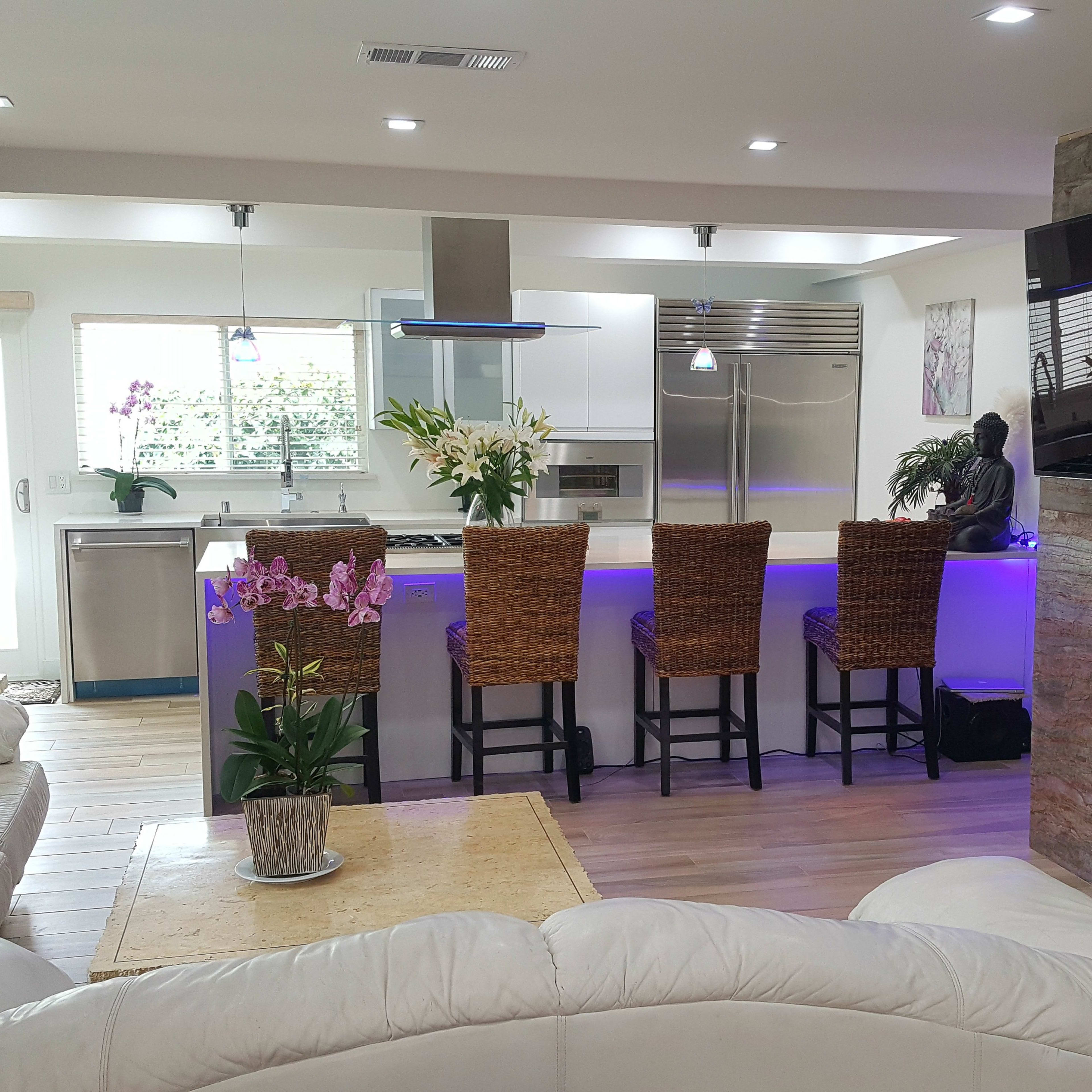 A modern living room with a white sectional sofa, a coffee table, and a kitchen area featuring bar stools, a large island, and a wall-mounted television above a stone fireplace.