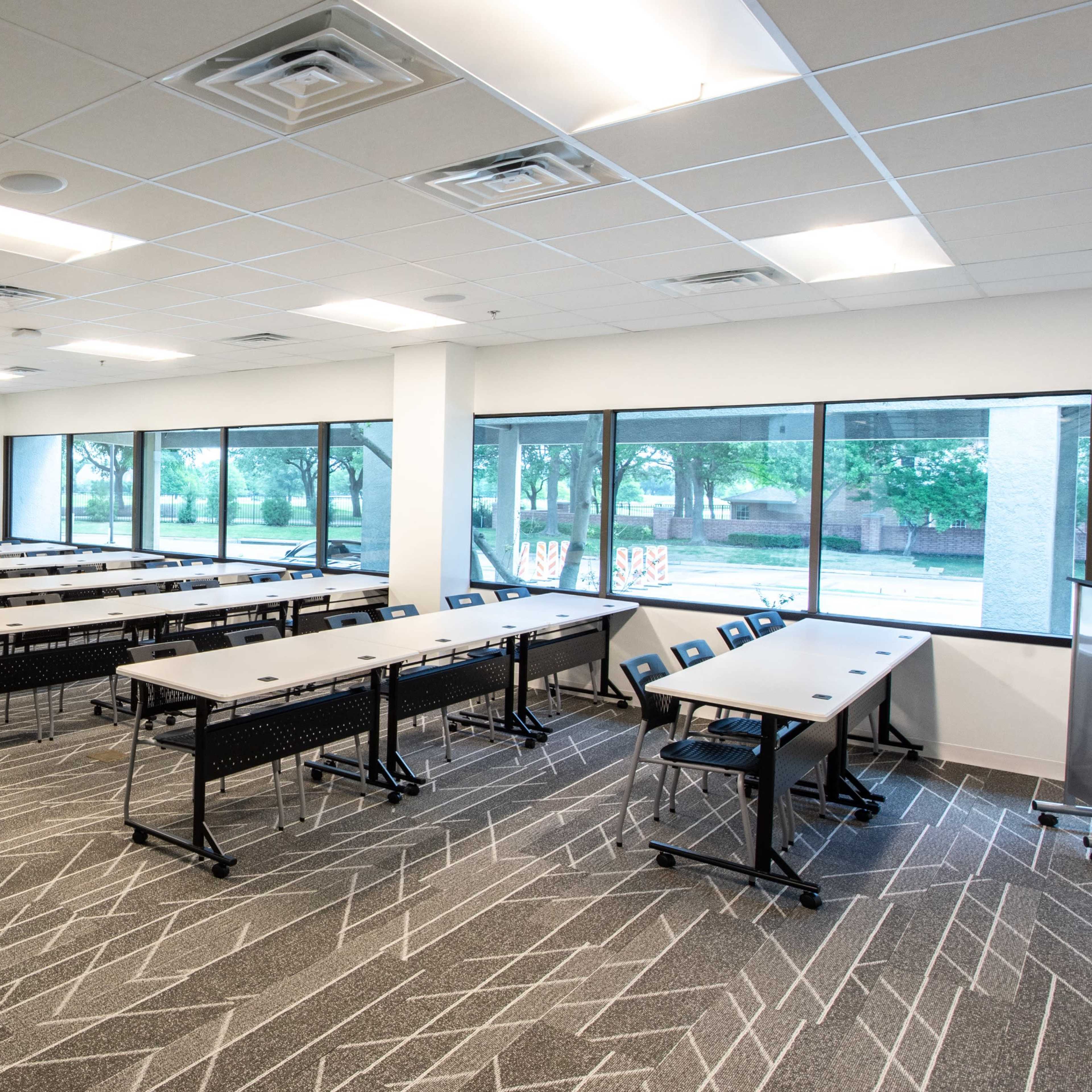 A spacious classroom arranged with several rows of tables and chairs, illuminated by large windows that provide natural light.