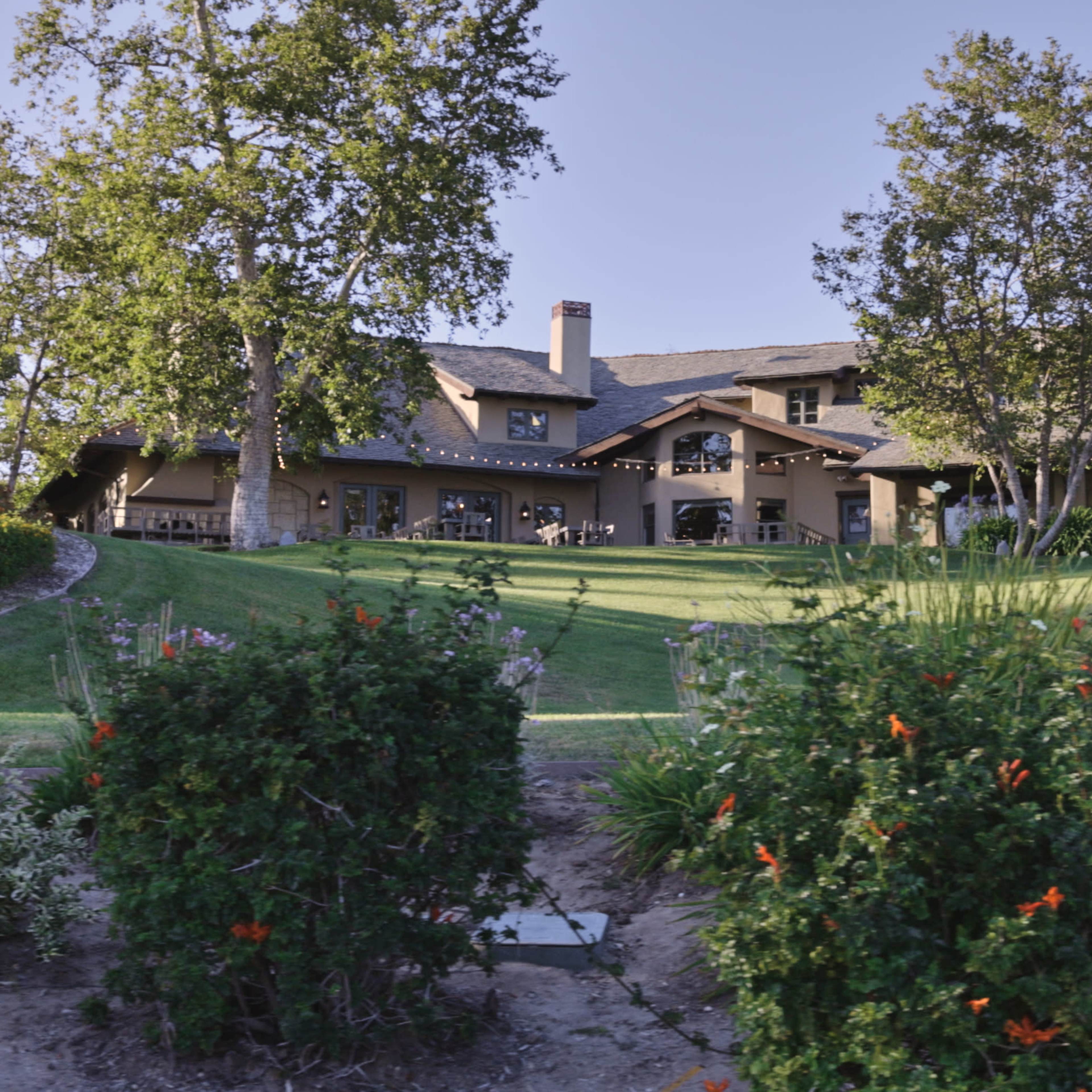 A large house with a sloped lawn, surrounded by trees and flower beds.