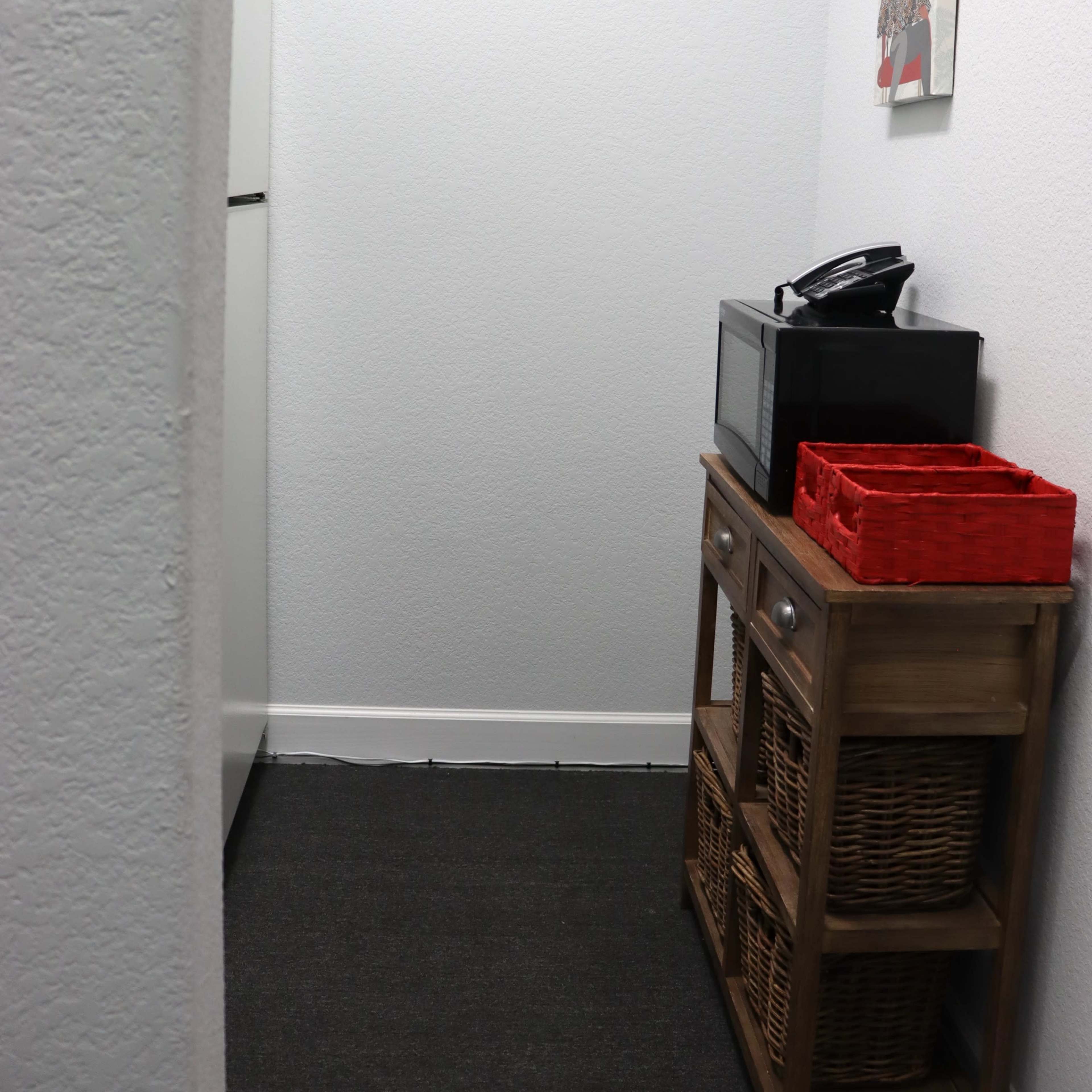 A narrow space features a refrigerator, a microwave on a wooden stand, and wicker baskets along with a framed picture on the wall.