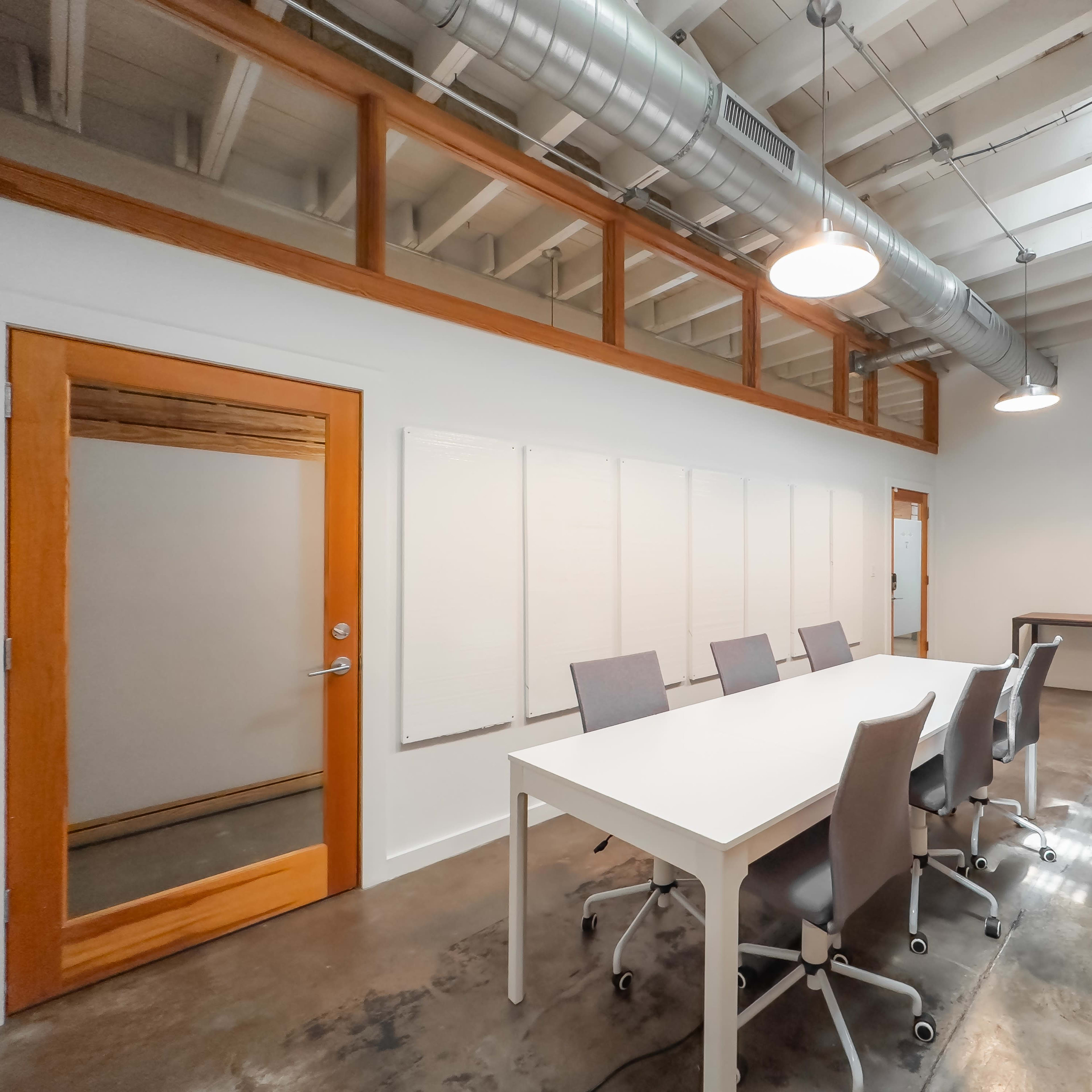 A modern conference room features a long white table surrounded by six gray chairs, with a glass-door meeting area and exposed ductwork overhead.
