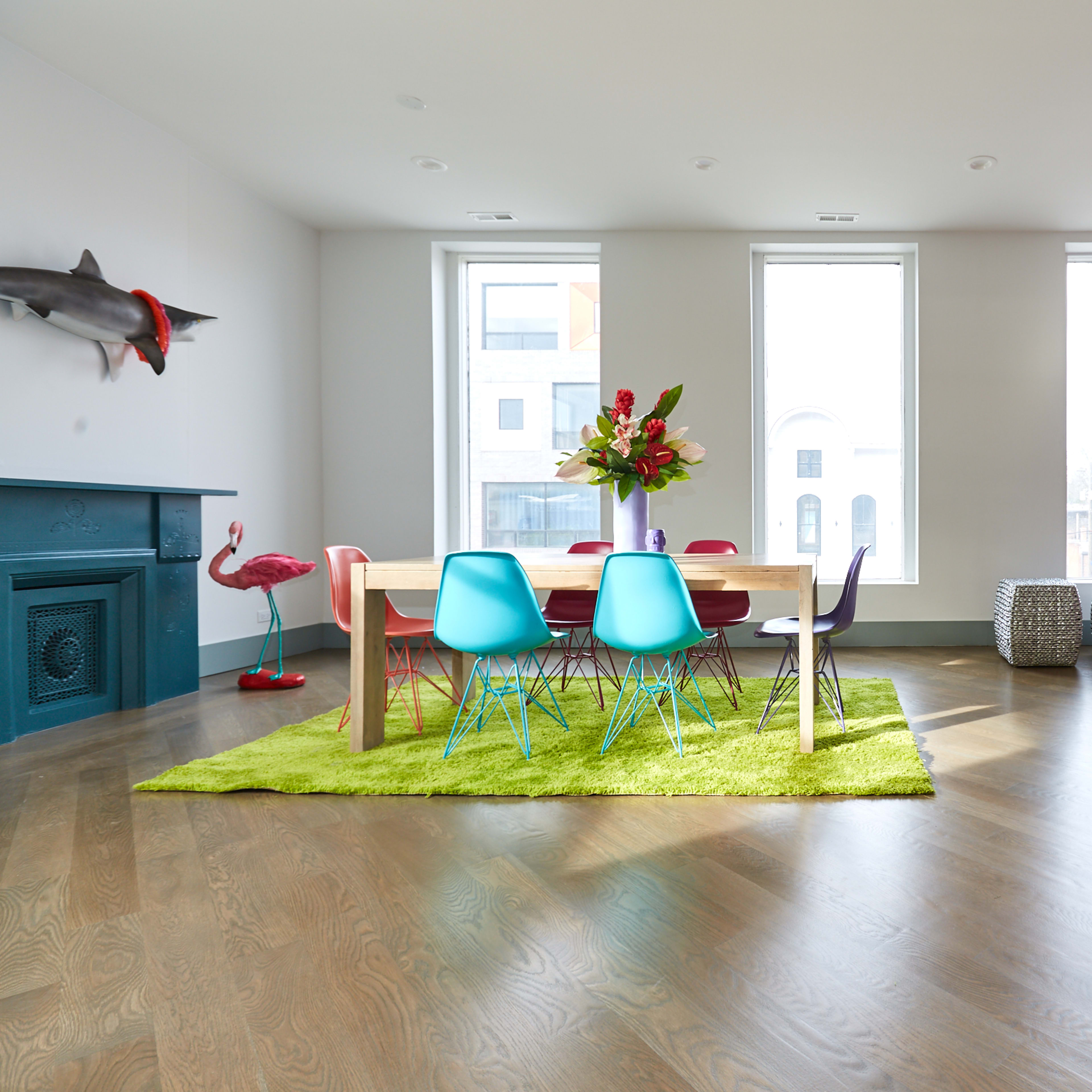 A brightly lit living space features a dining table surrounded by colorful chairs on a green rug, with large windows and decorative elements including a flamingo statue and fish mounts on the walls.