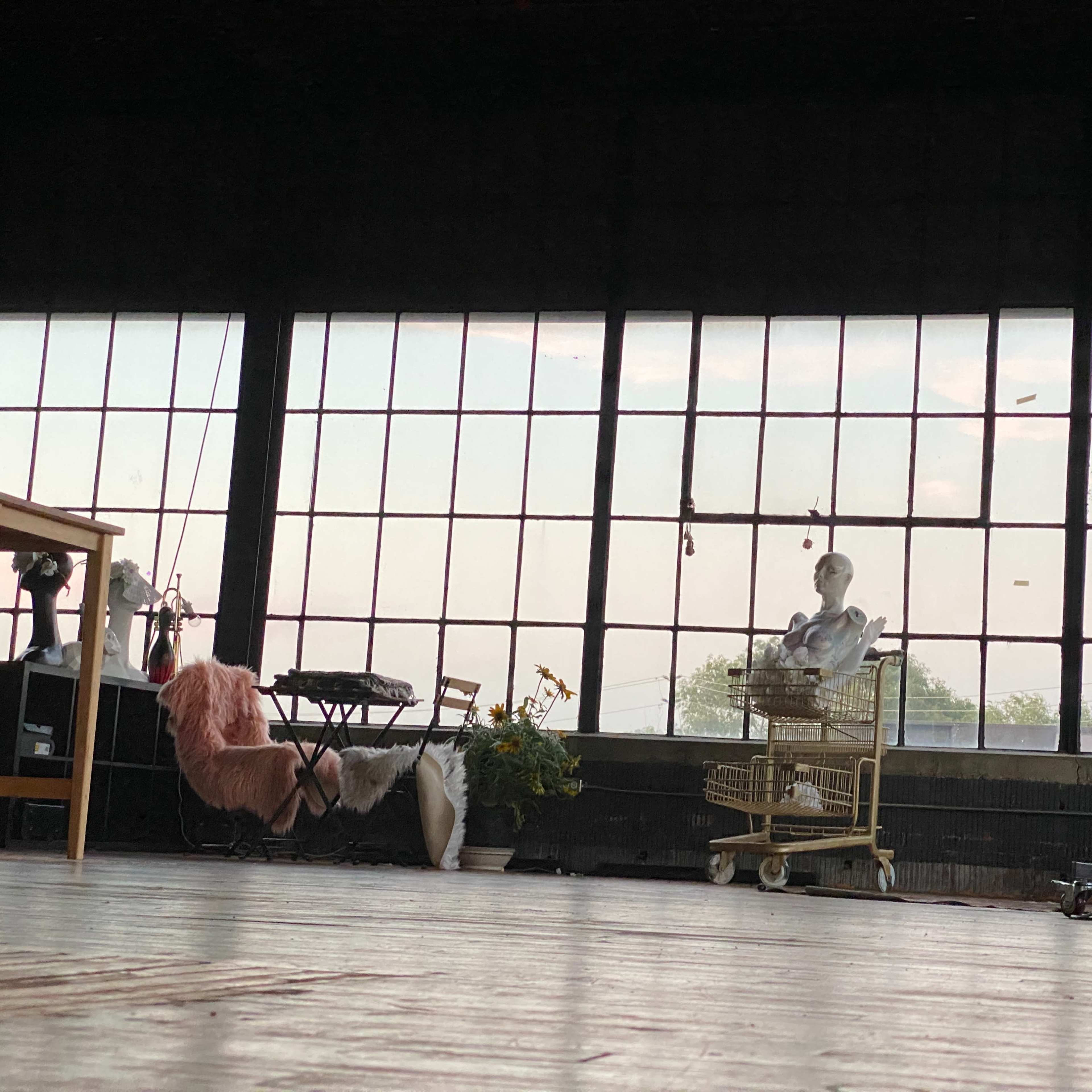 A spacious room with large windows, a mannequin in a shopping cart, a plant, and various photography equipment.