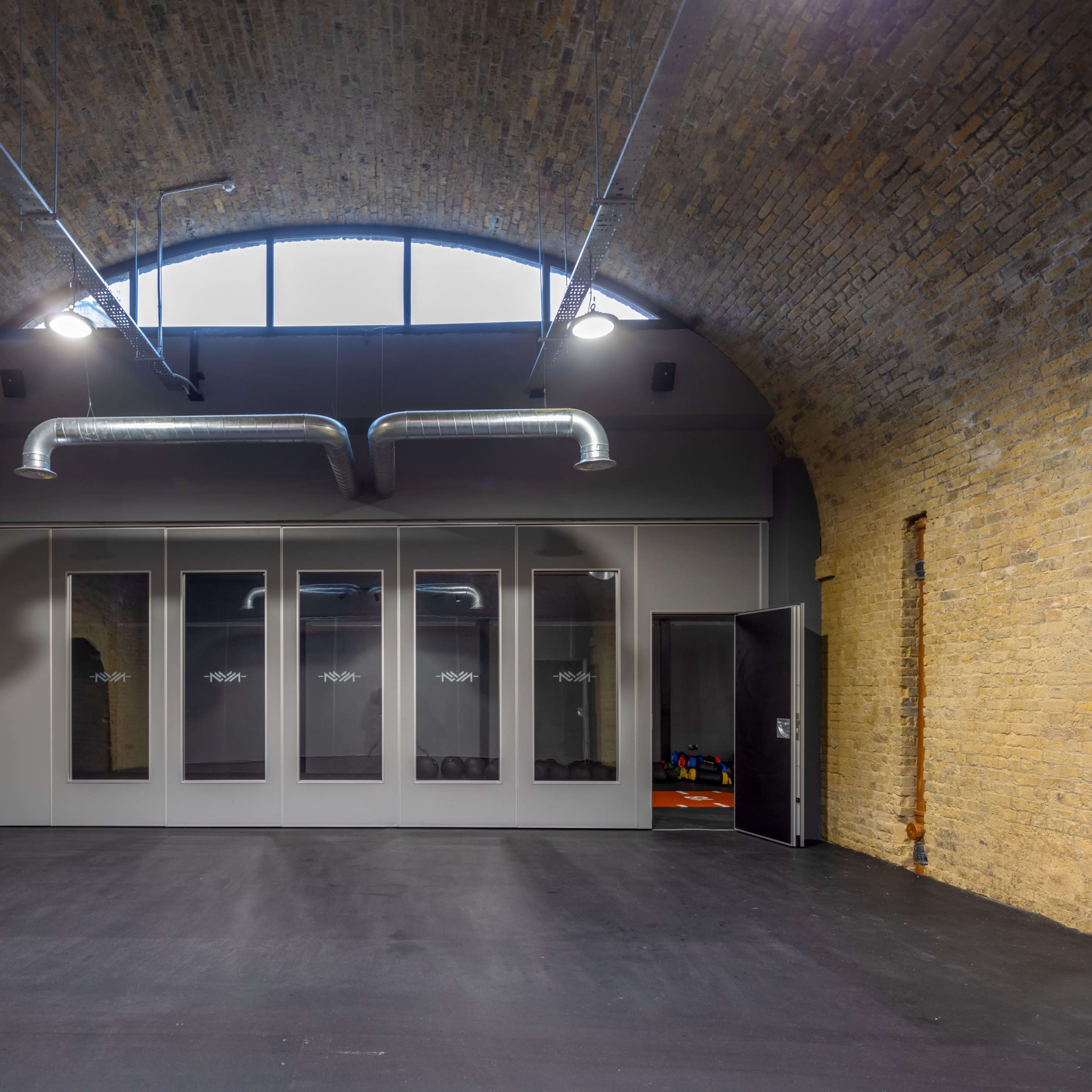 An interior gym space with exposed brick walls, large windows, and a mirrored wall with an entrance to a room containing colorful exercise equipment.