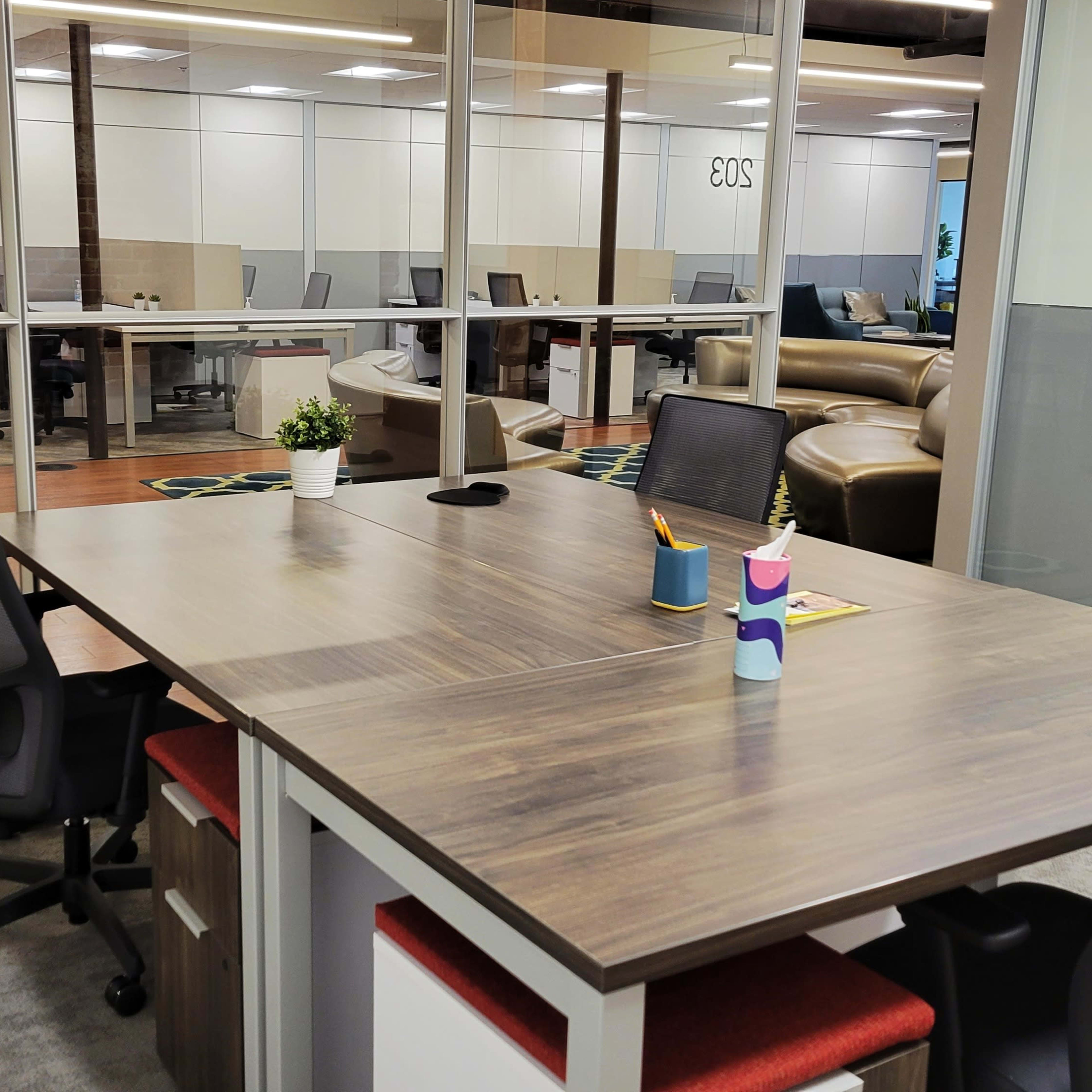 A modern office space with a large wooden table at the forefront, flanked by two black chairs and surrounded by glass-walled workstations in the background.