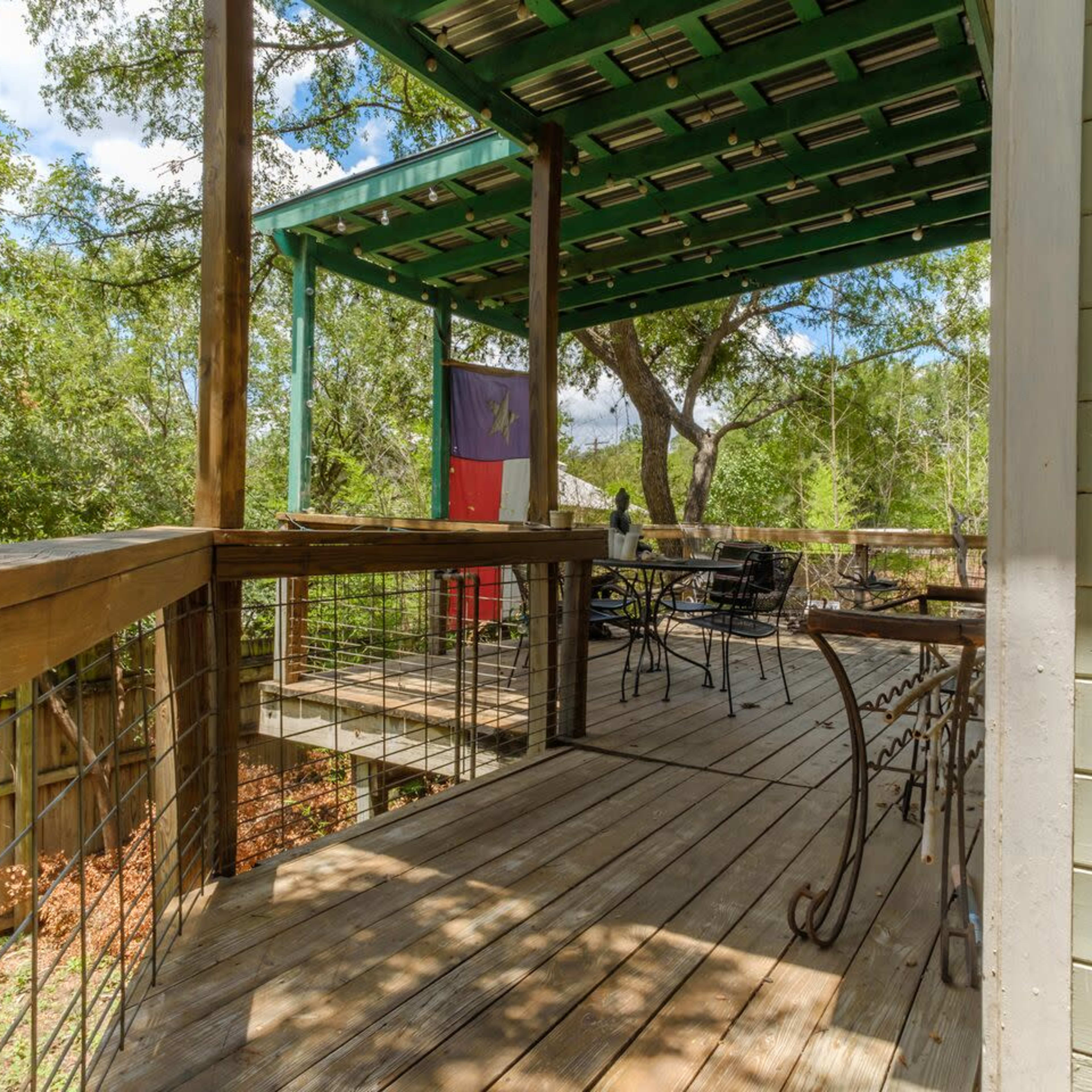 A wooden deck with a green roof, featuring a flag and outdoor furniture surrounded by trees.