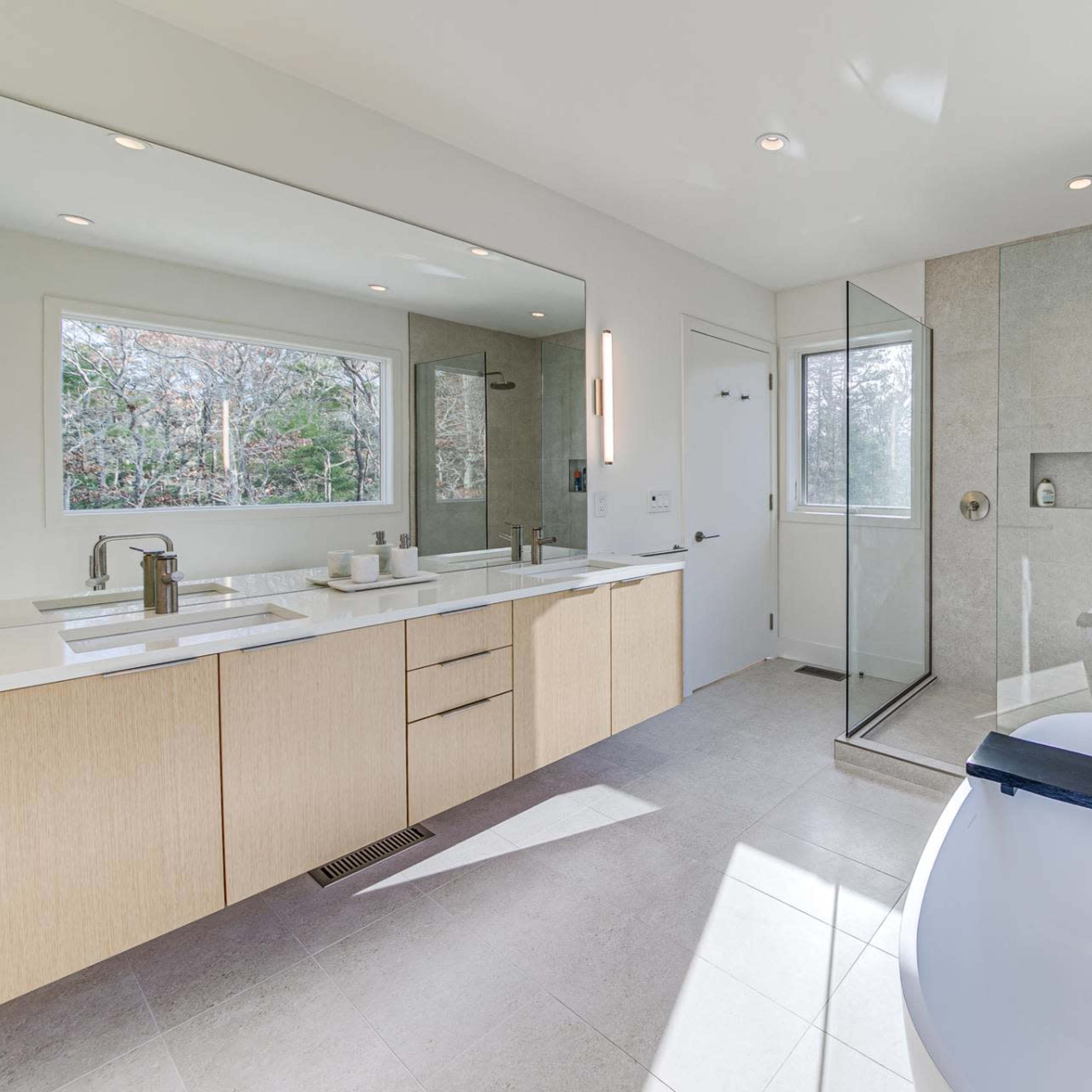 A modern bathroom featuring a large mirror, a double vanity with wooden cabinetry, a glass shower enclosure, and a freestanding bathtub beside a window.