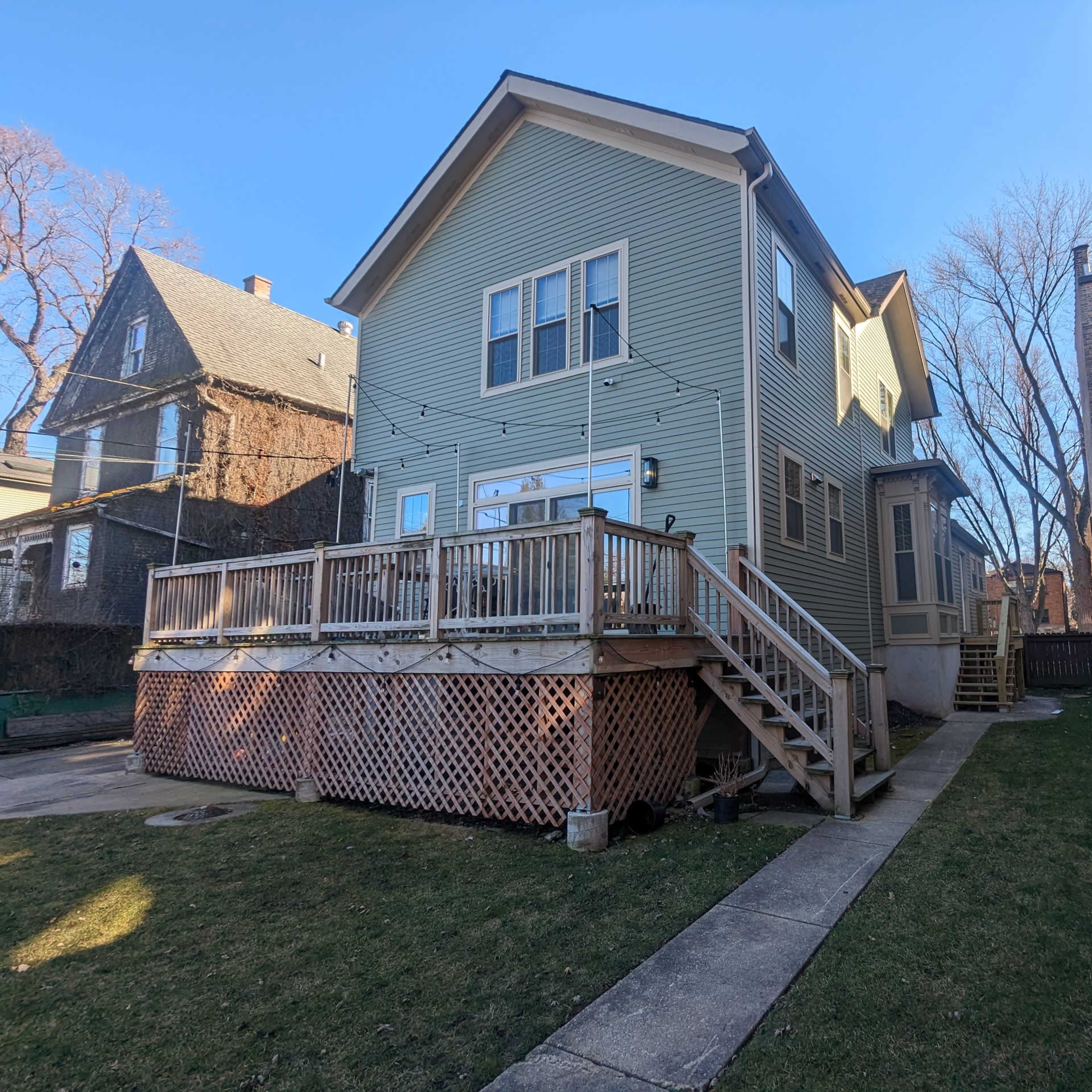 A two-story green house with a wooden deck on one side, situated alongside a narrow path and adjacent to a brick building.