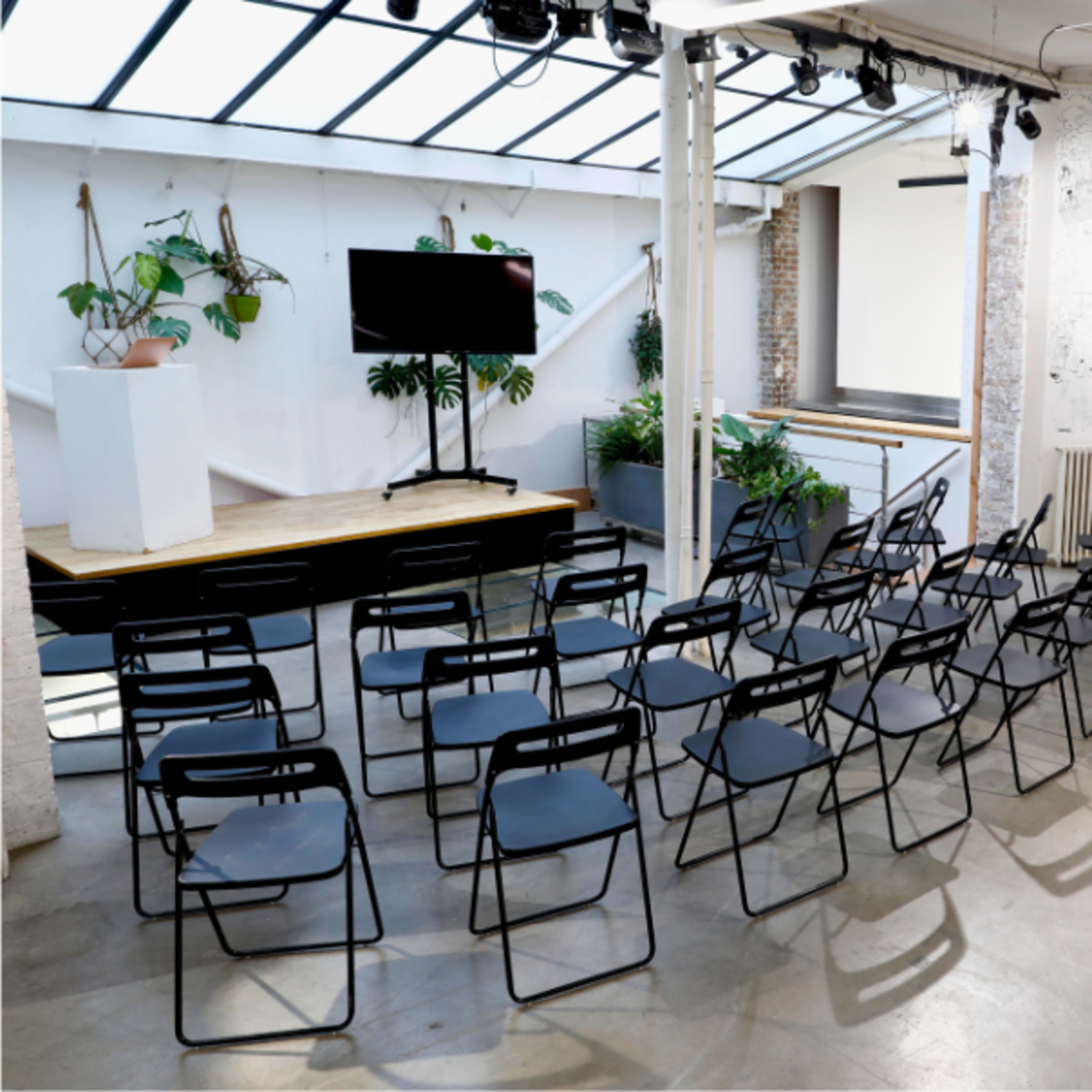 A modern event space with arranged black folding chairs facing a small stage and a large TV screen, surrounded by potted plants.