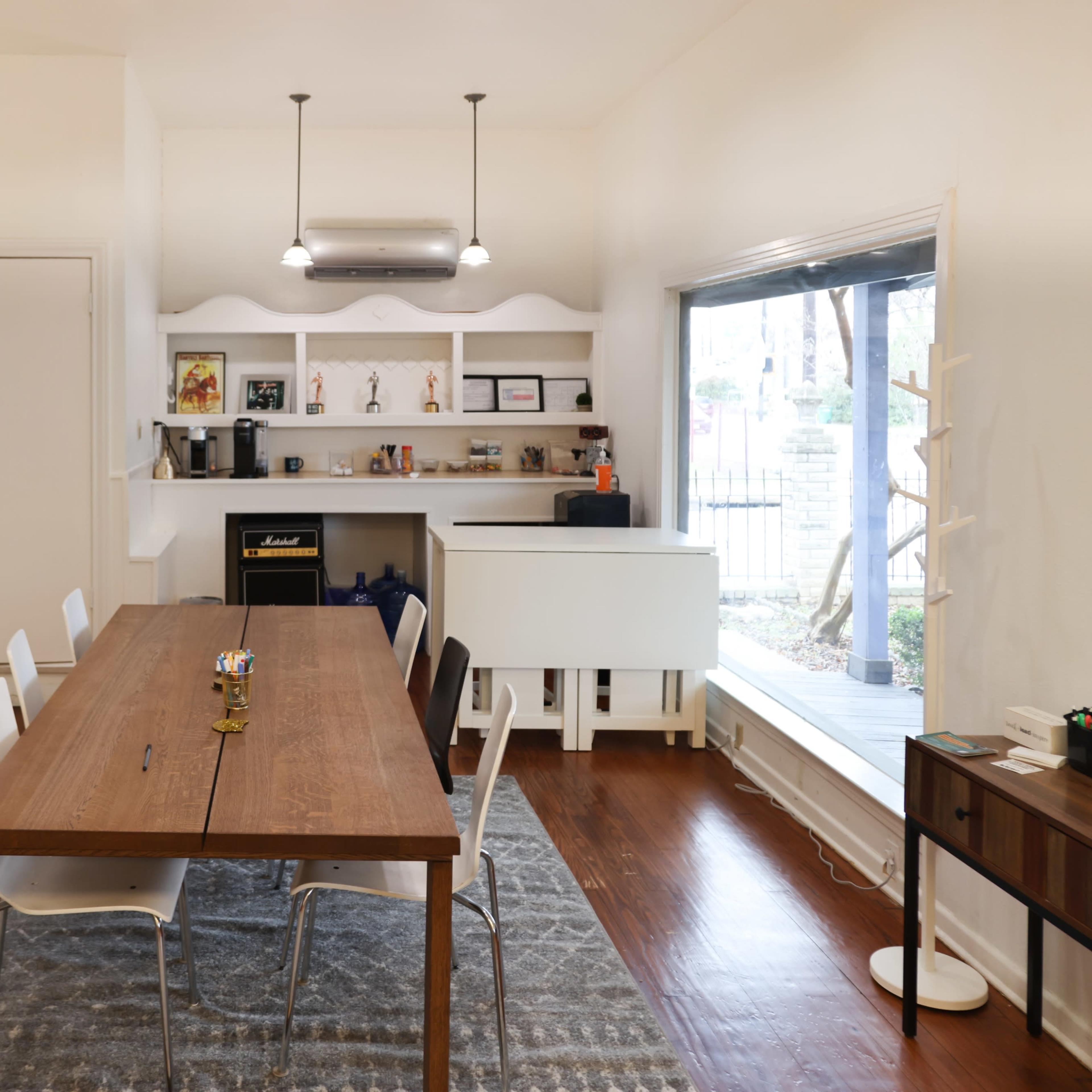A modern dining area features a wooden table surrounded by white chairs, with a small kitchen counter and a window overlooking an outdoor space.