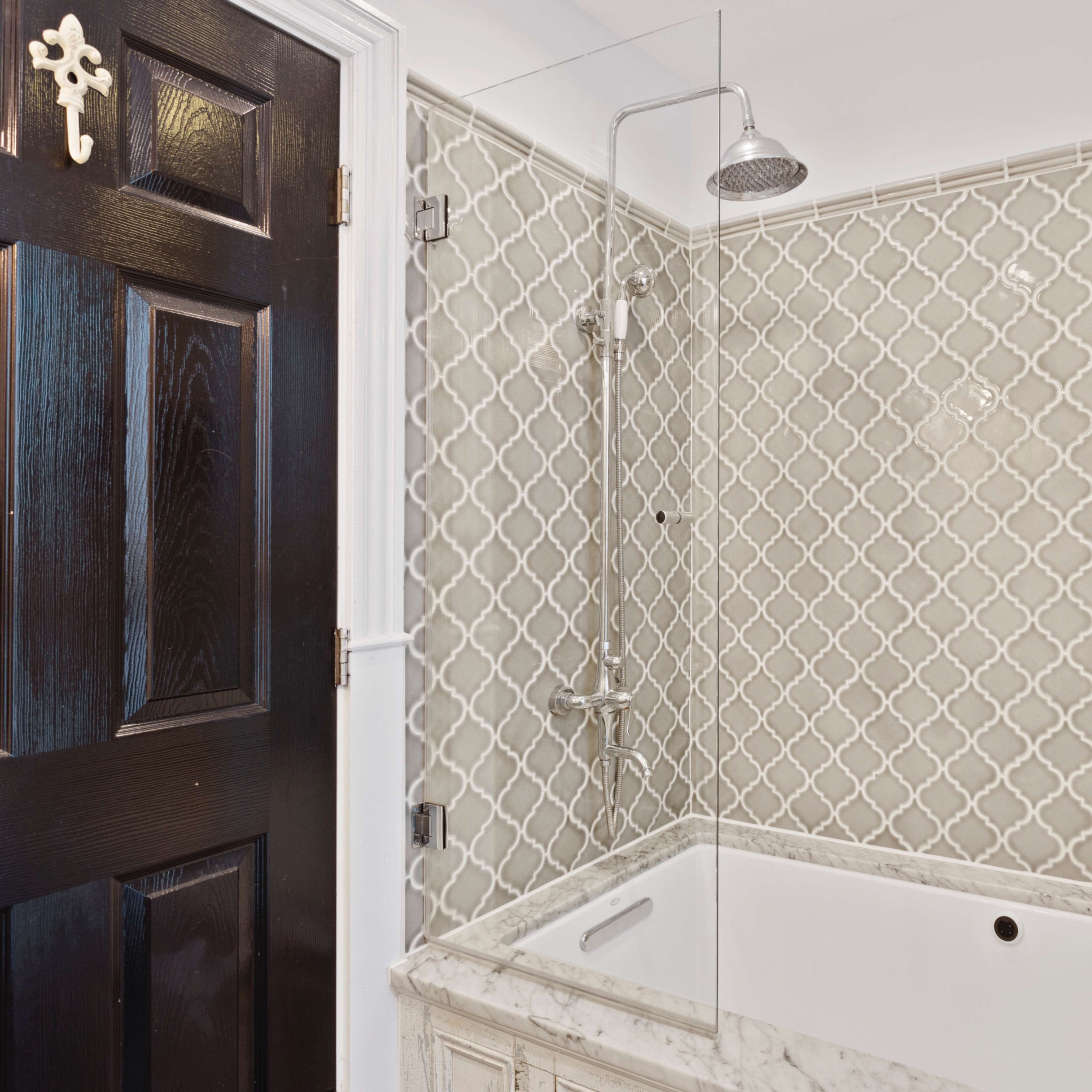 A modern bathroom with a glass shower enclosure, a tiled wall featuring a geometric pattern, and a dark wooden door.