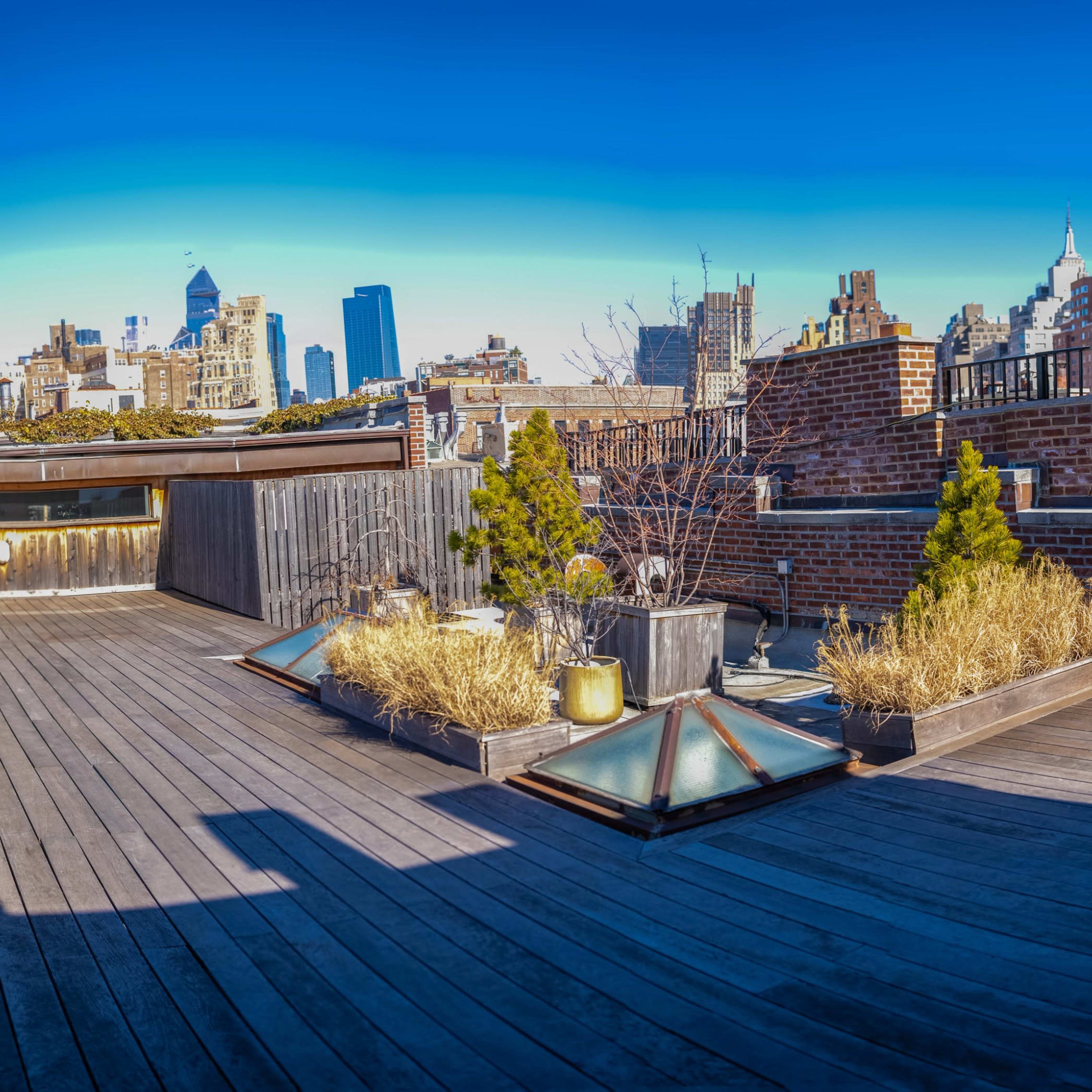 A spacious rooftop terrace featuring wooden flooring, potted plants, and a city skyline in the background under a clear blue sky.