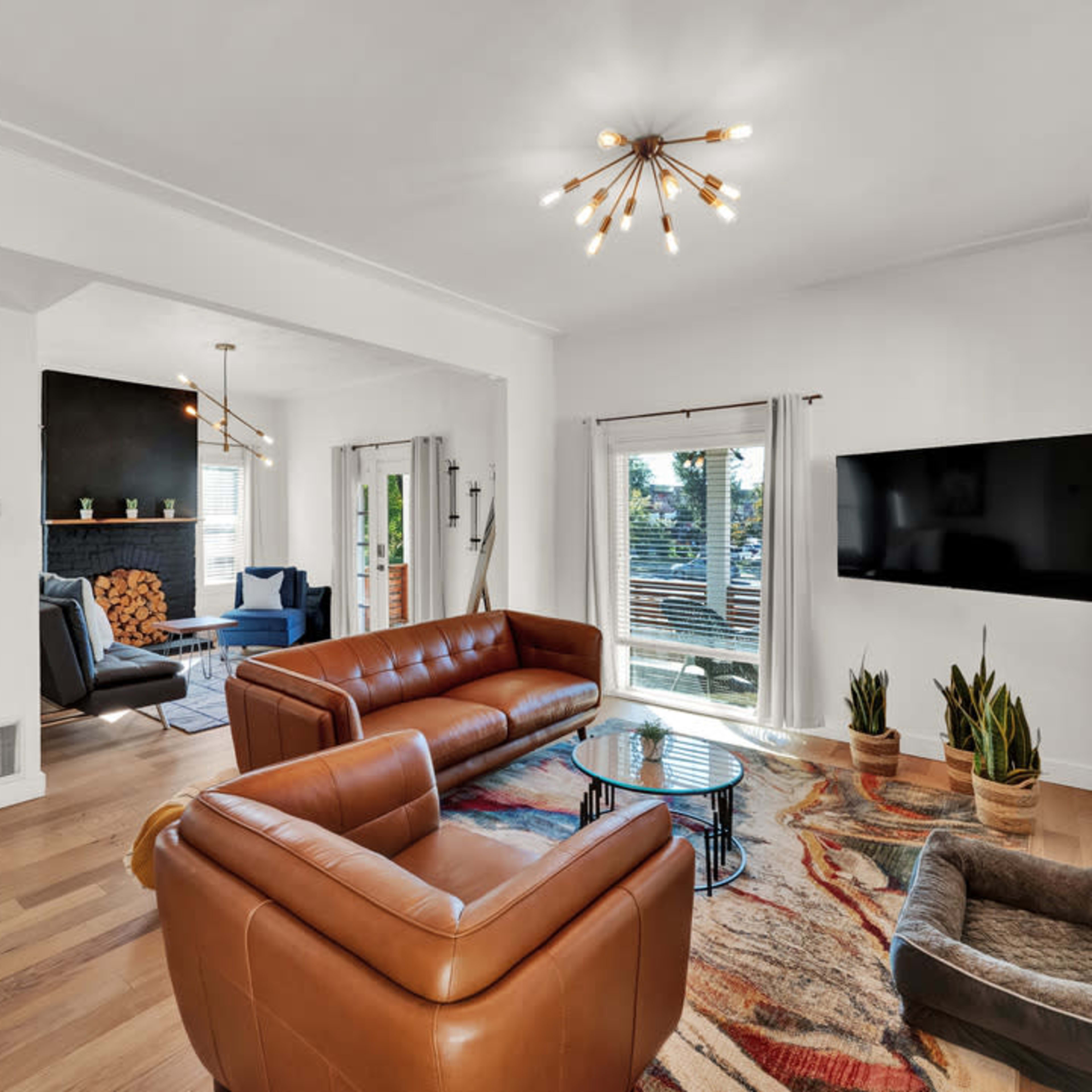 A cozy living room features brown leather sofas, a circular coffee table, a mounted television, and large windows that provide natural light.