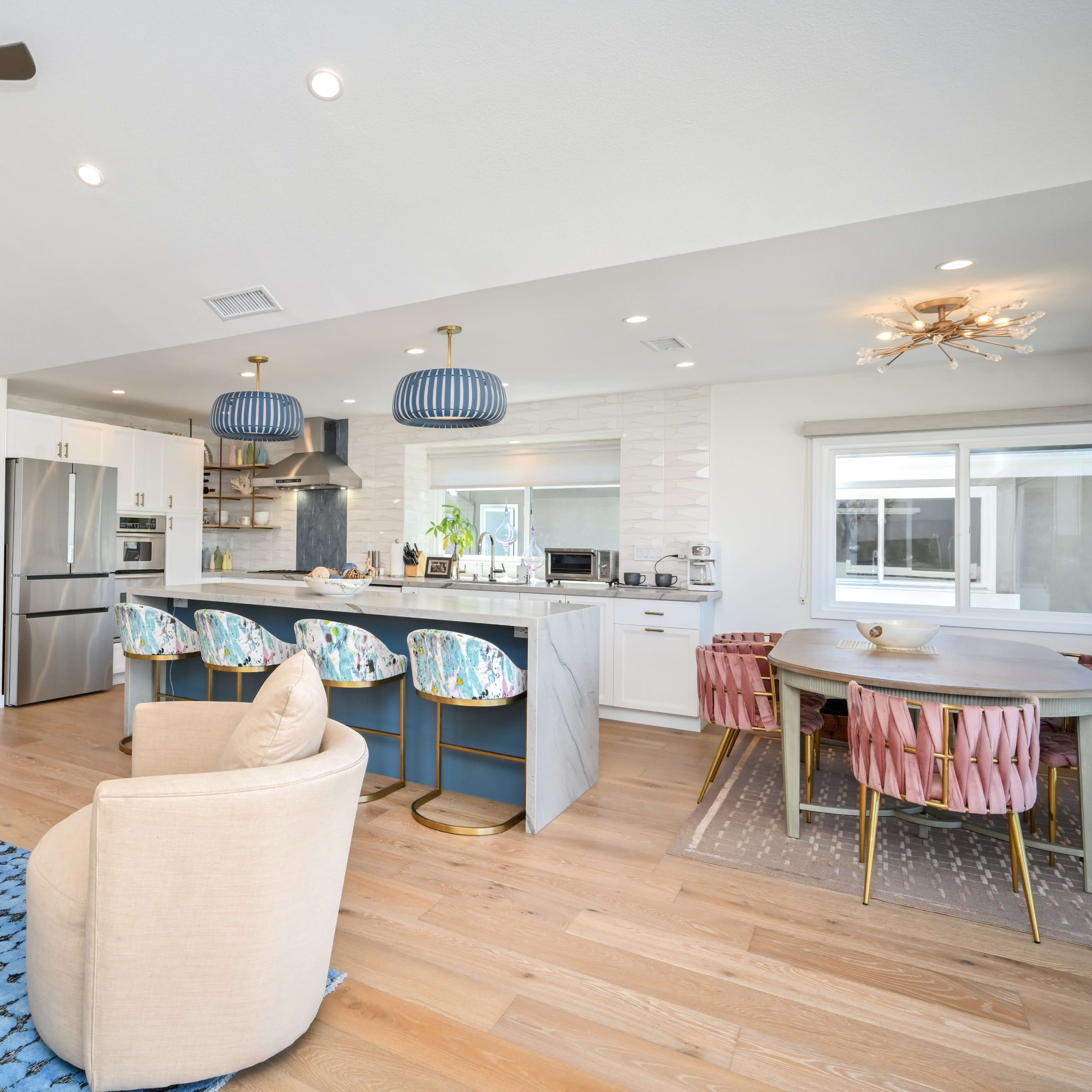 A modern open-concept kitchen and living area features sleek appliances, pastel-colored bar stools, and a dining table with pink chairs.