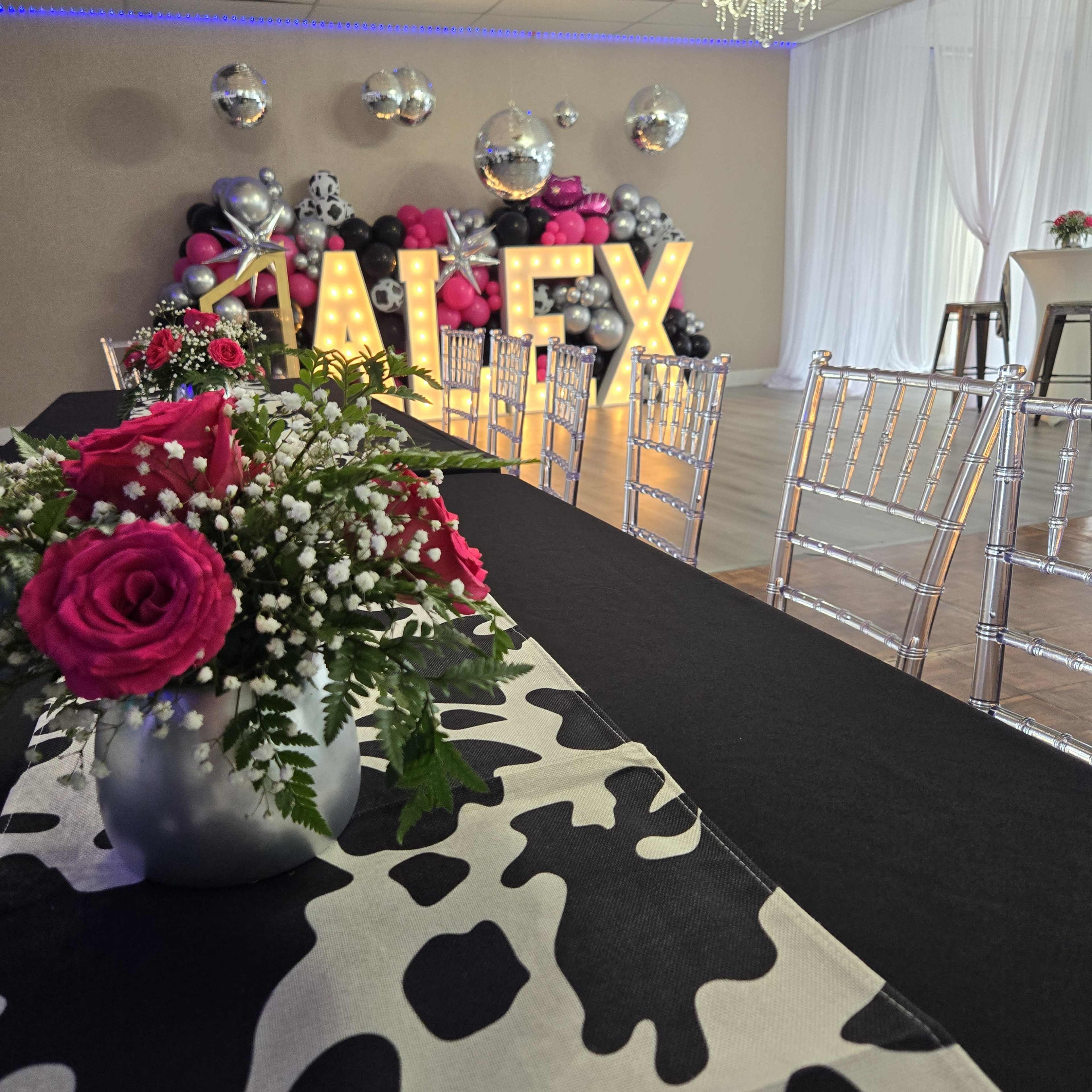 A decorated event space with a long table covered in a cow print table runner, floral arrangements, and large illuminated letters spelling "ALEX" against a backdrop of balloons and disco balls.
