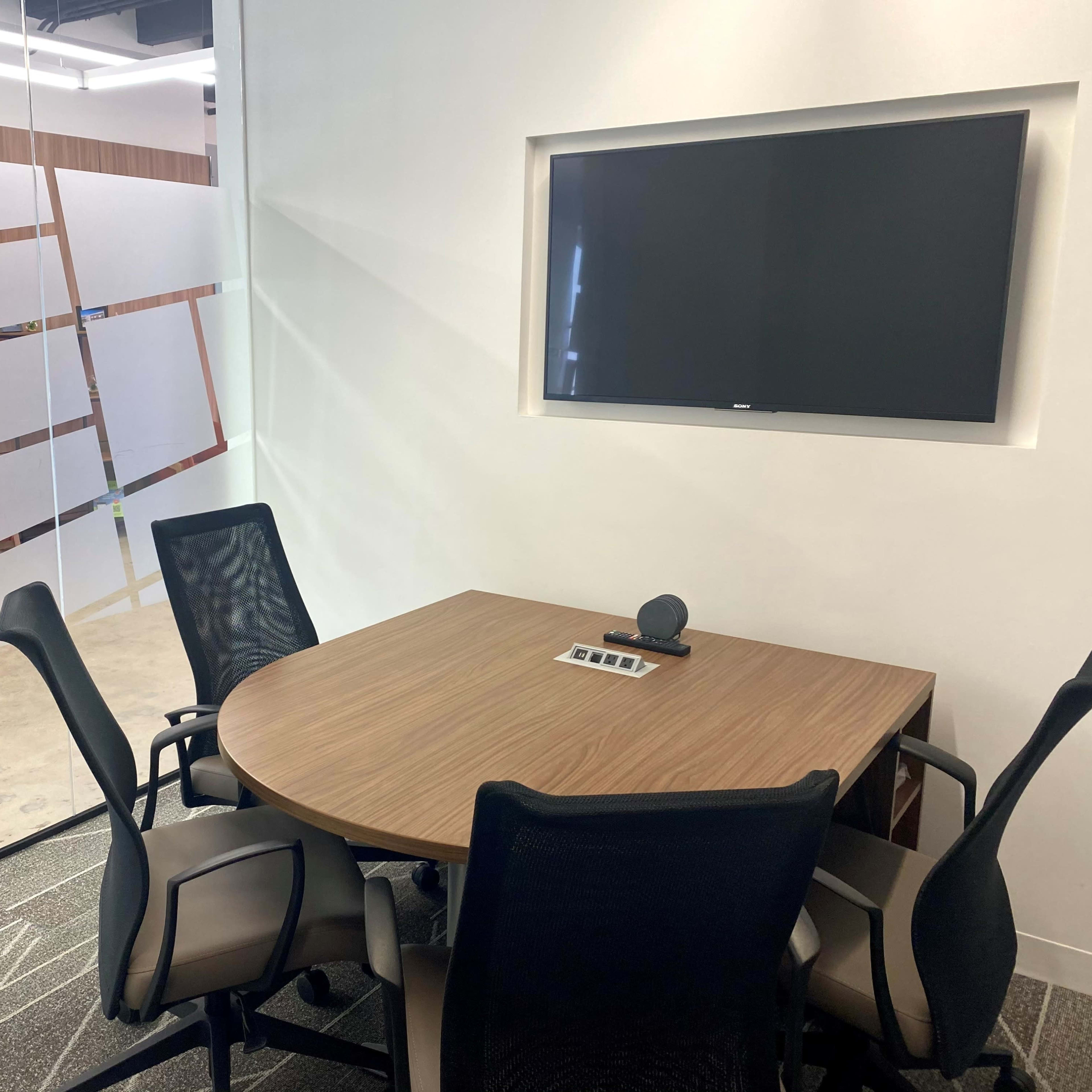 A small conference room features a round wooden table, five chairs, a wall-mounted television, and a glass panel with geometric designs.