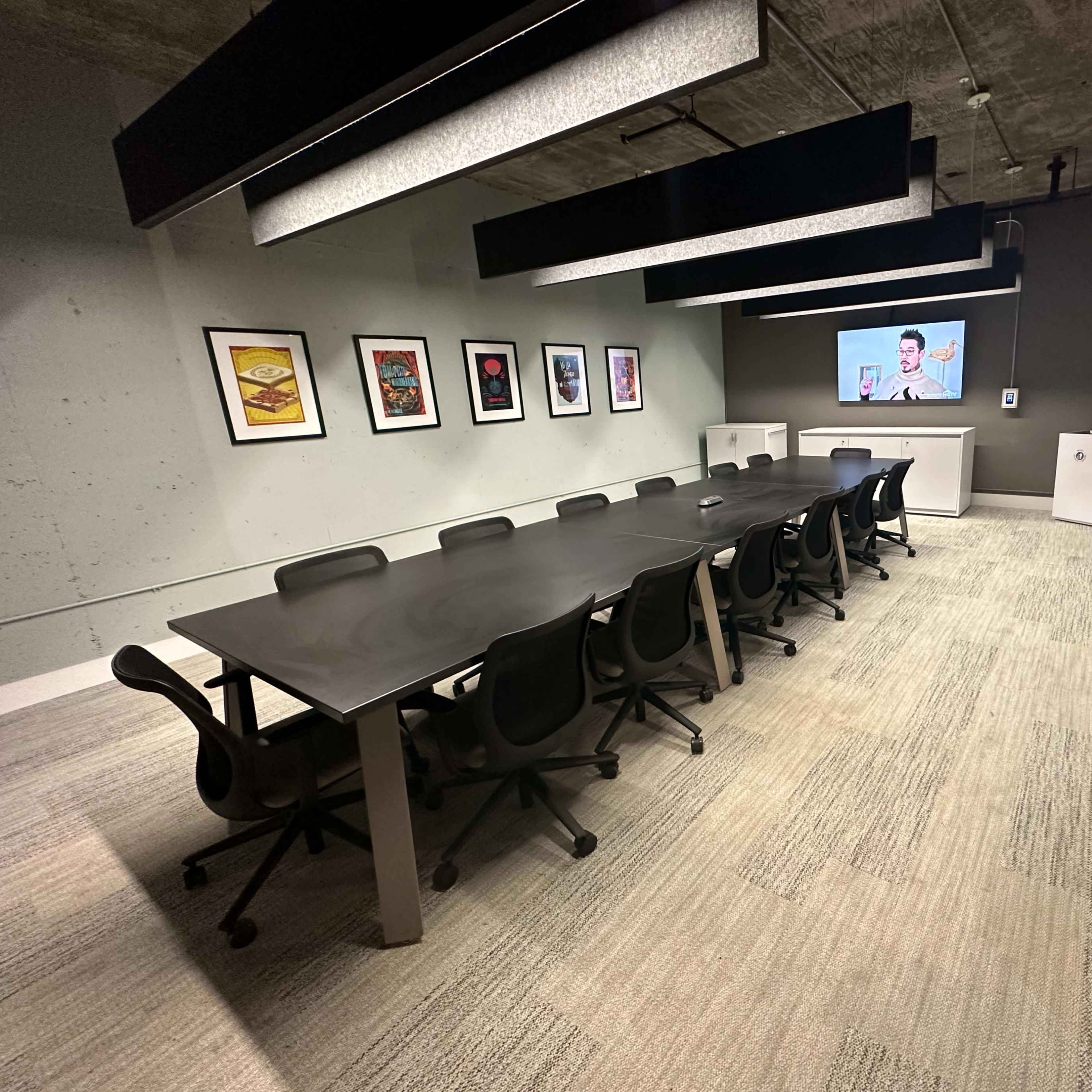 A long rectangular conference table is set up in a modern meeting room with surround black chairs, artwork on the walls, and a presentation screen at one end.