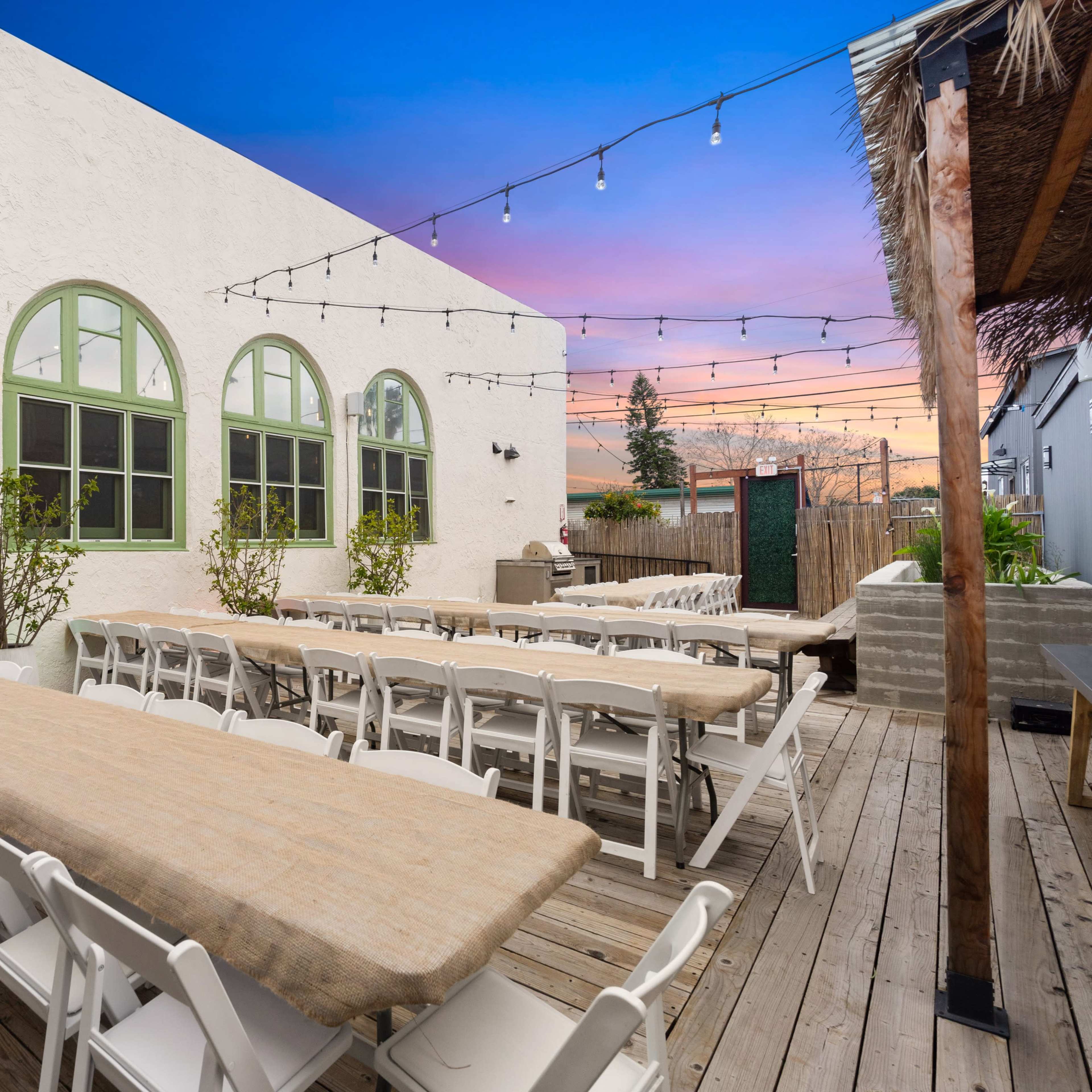 A patio area with long tables set up for dining, lined by greenery and illuminated by string lights against a colorful sunset sky.