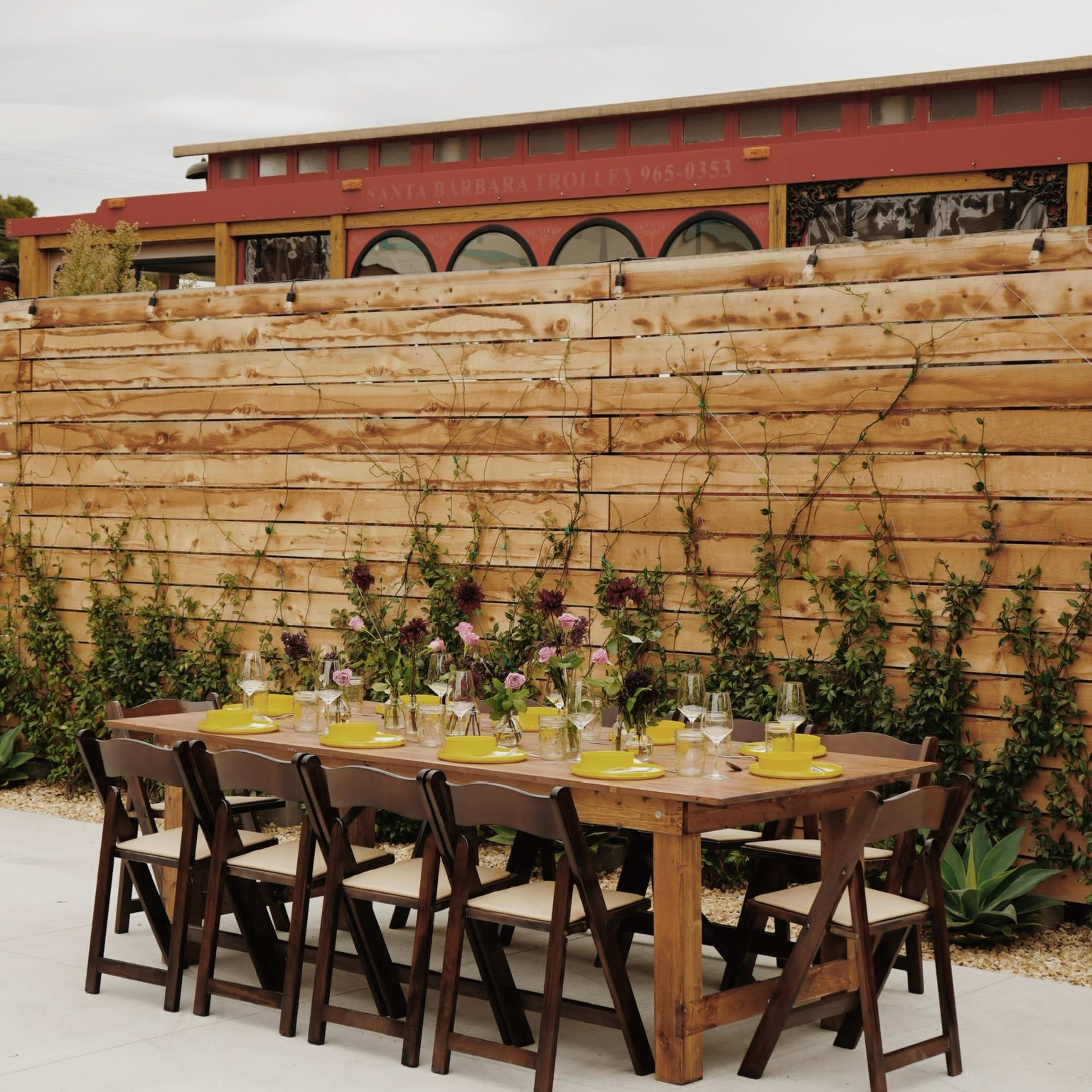 A long wooden dining table is set with decorative plates and flowers against a backdrop of a wooden fence and a red building.