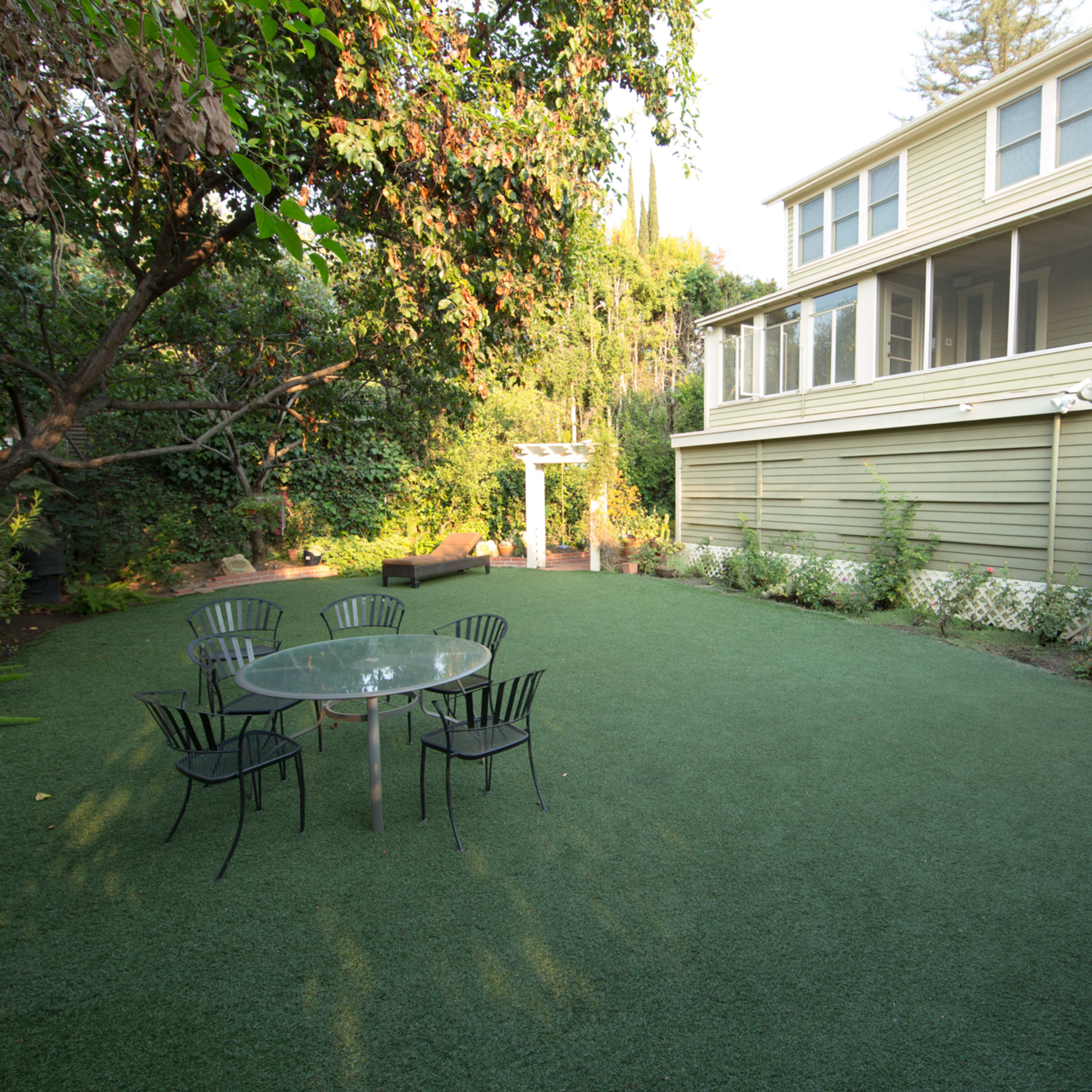 A backyard with artificial grass, featuring a glass table surrounded by black chairs and a multi-story house in the background.