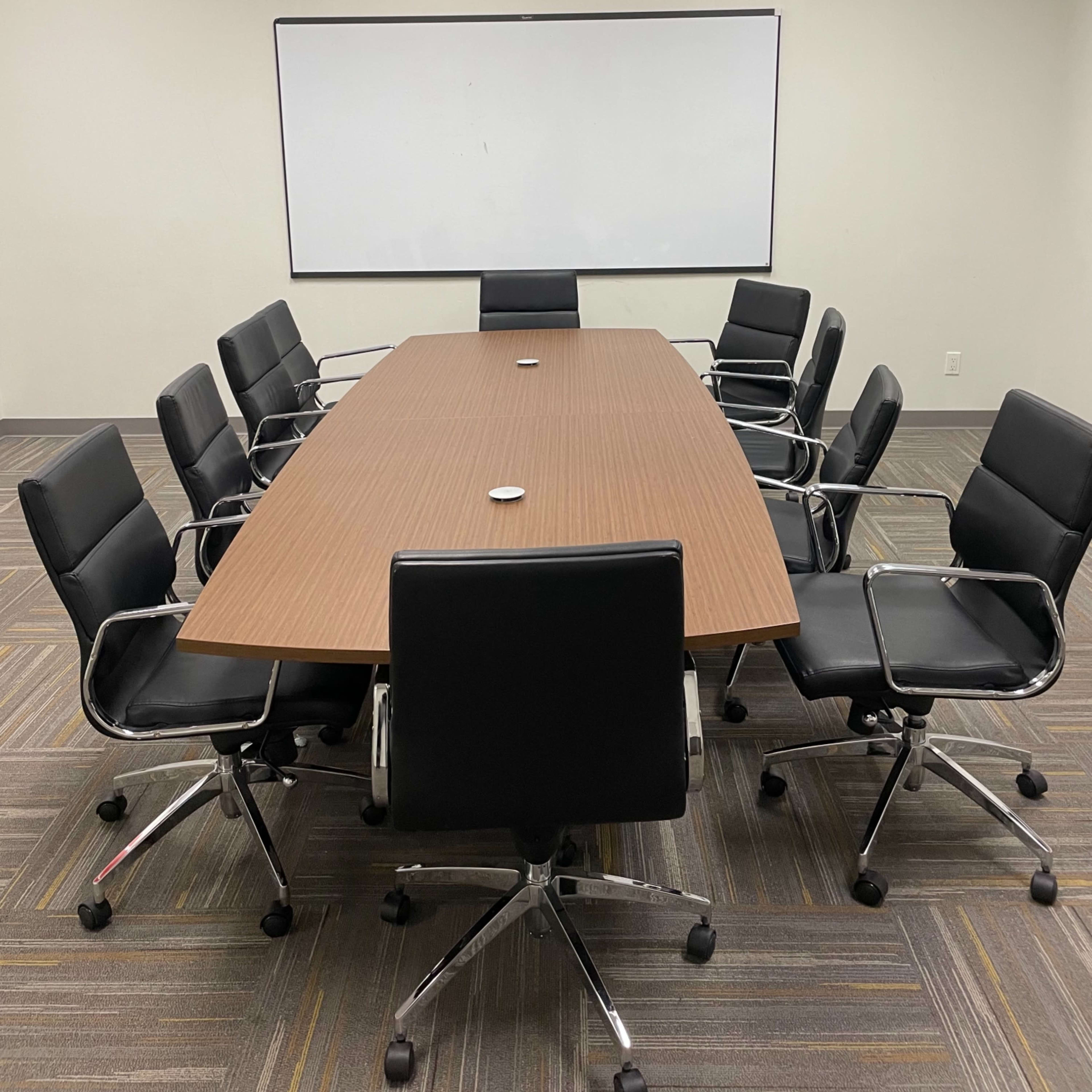 A conference room features a long wooden table surrounded by eight black rolling chairs and a whiteboard on the wall.