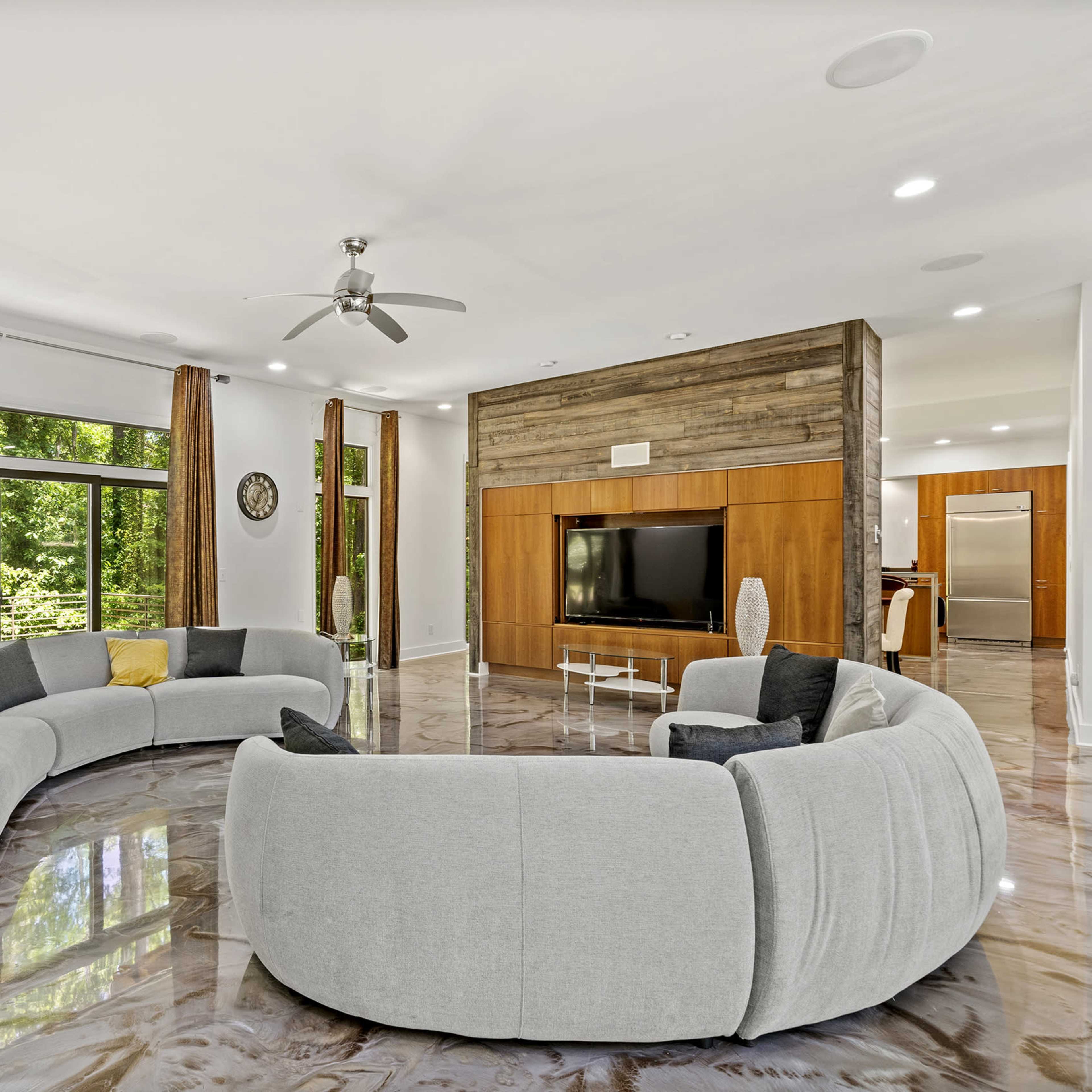 A modern living room featuring a curved gray sofa, large windows with natural light, and a wooden accent wall housing a television.