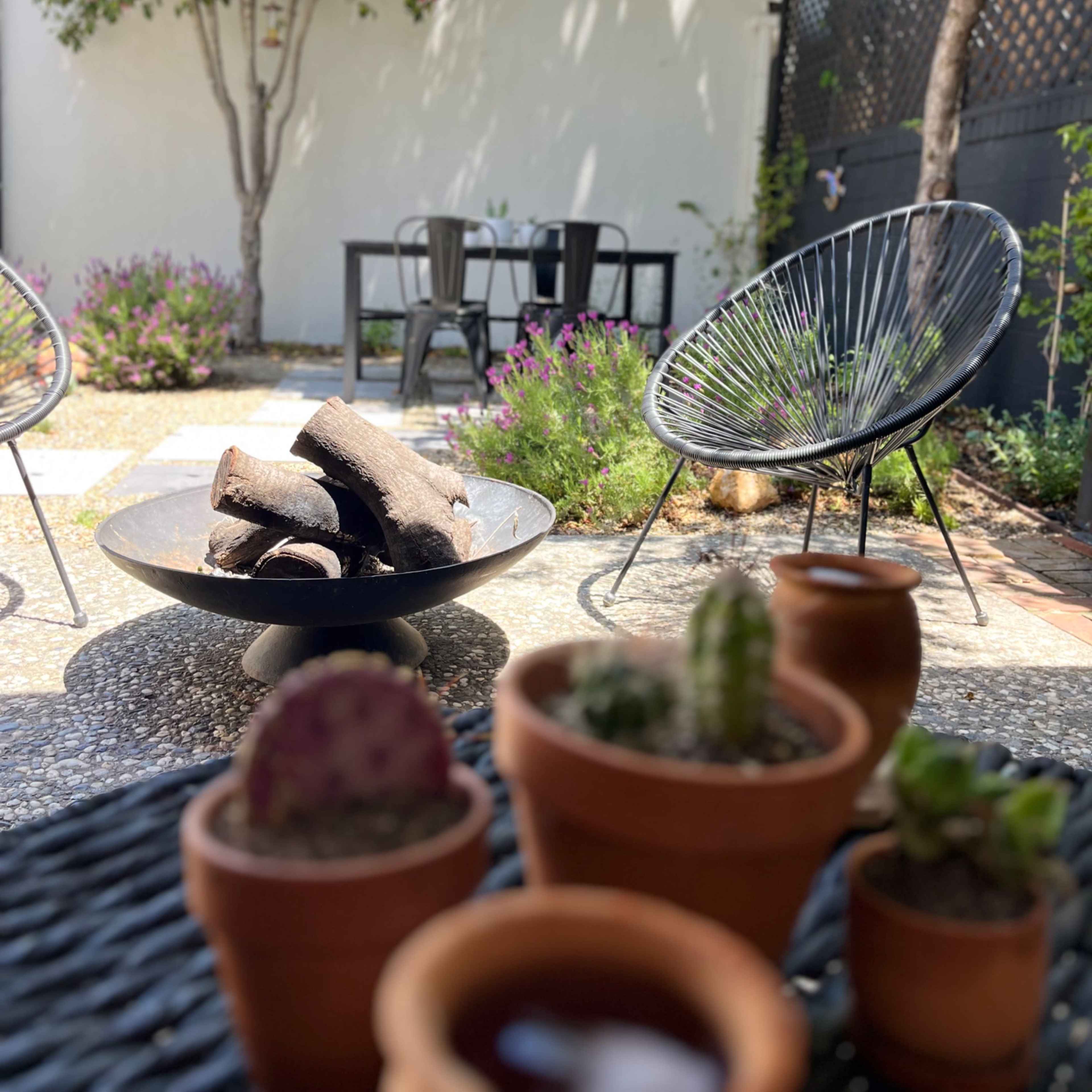 A patio scene features a fire pit surrounded by modern chairs, with a table in the background and potted plants in the foreground.