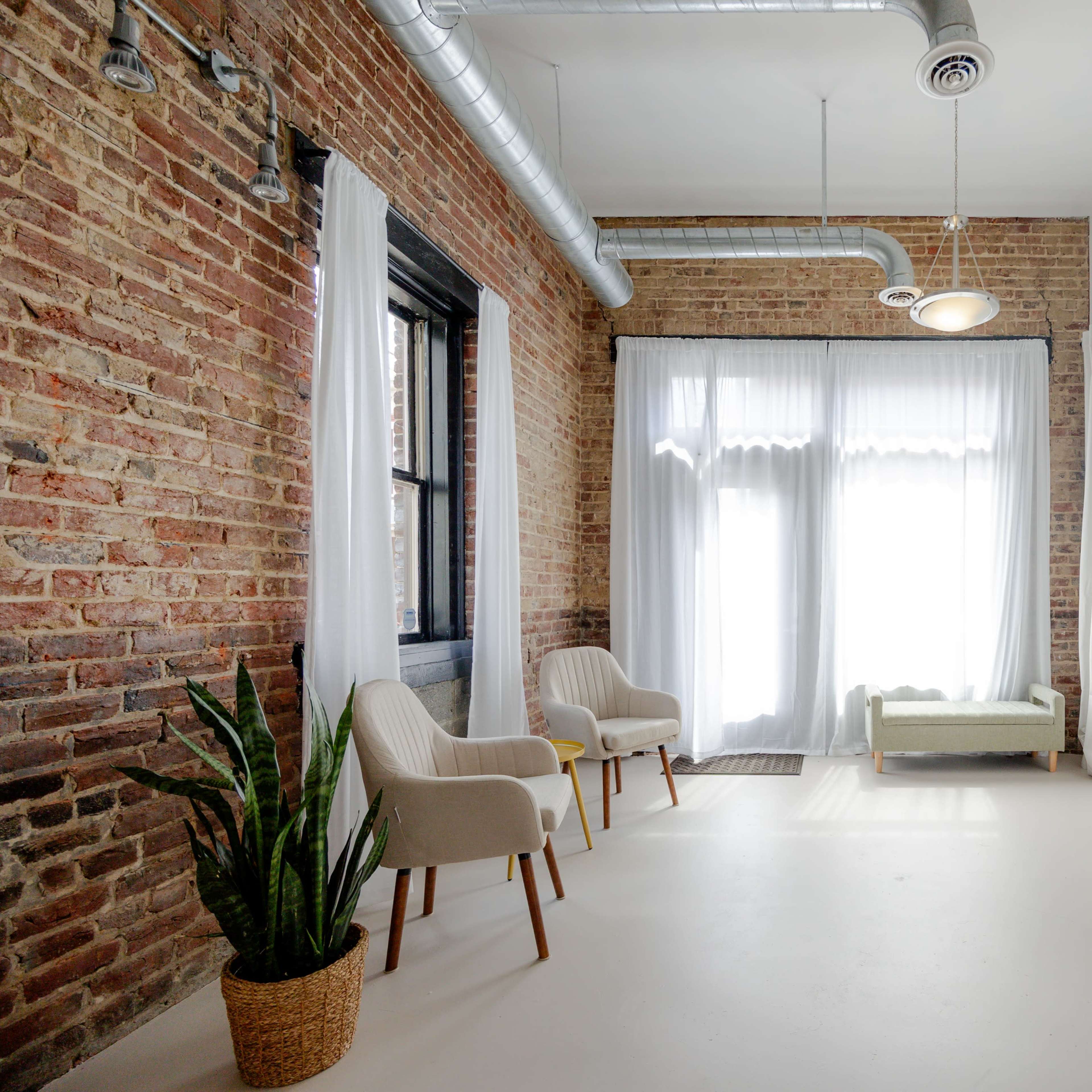 A spacious room with exposed brick walls, large windows covered by white curtains, two upholstered chairs, and a potted plant in the corner.