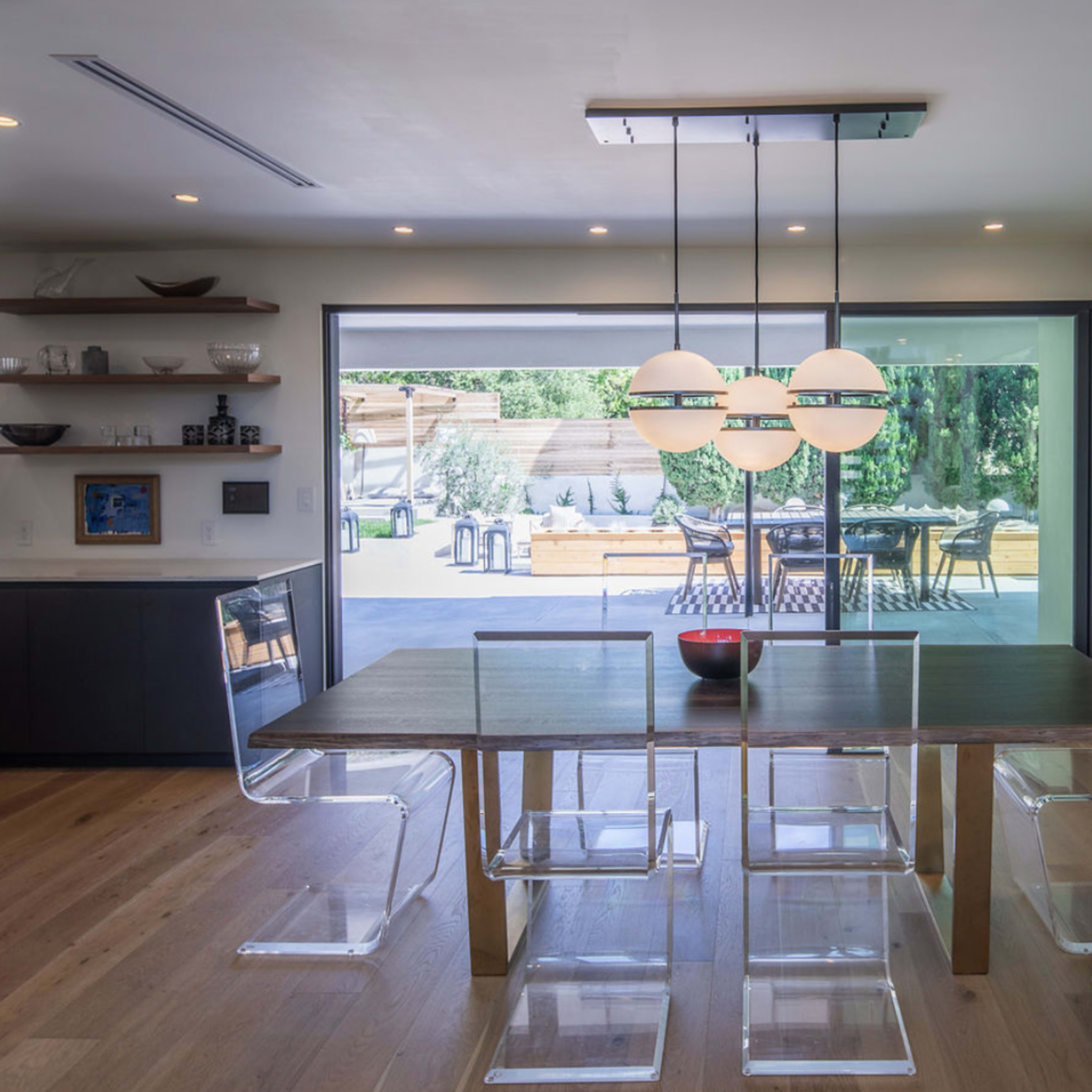 A modern dining area features a wooden table surrounded by transparent chairs, with a view of an outdoor patio through large glass doors.