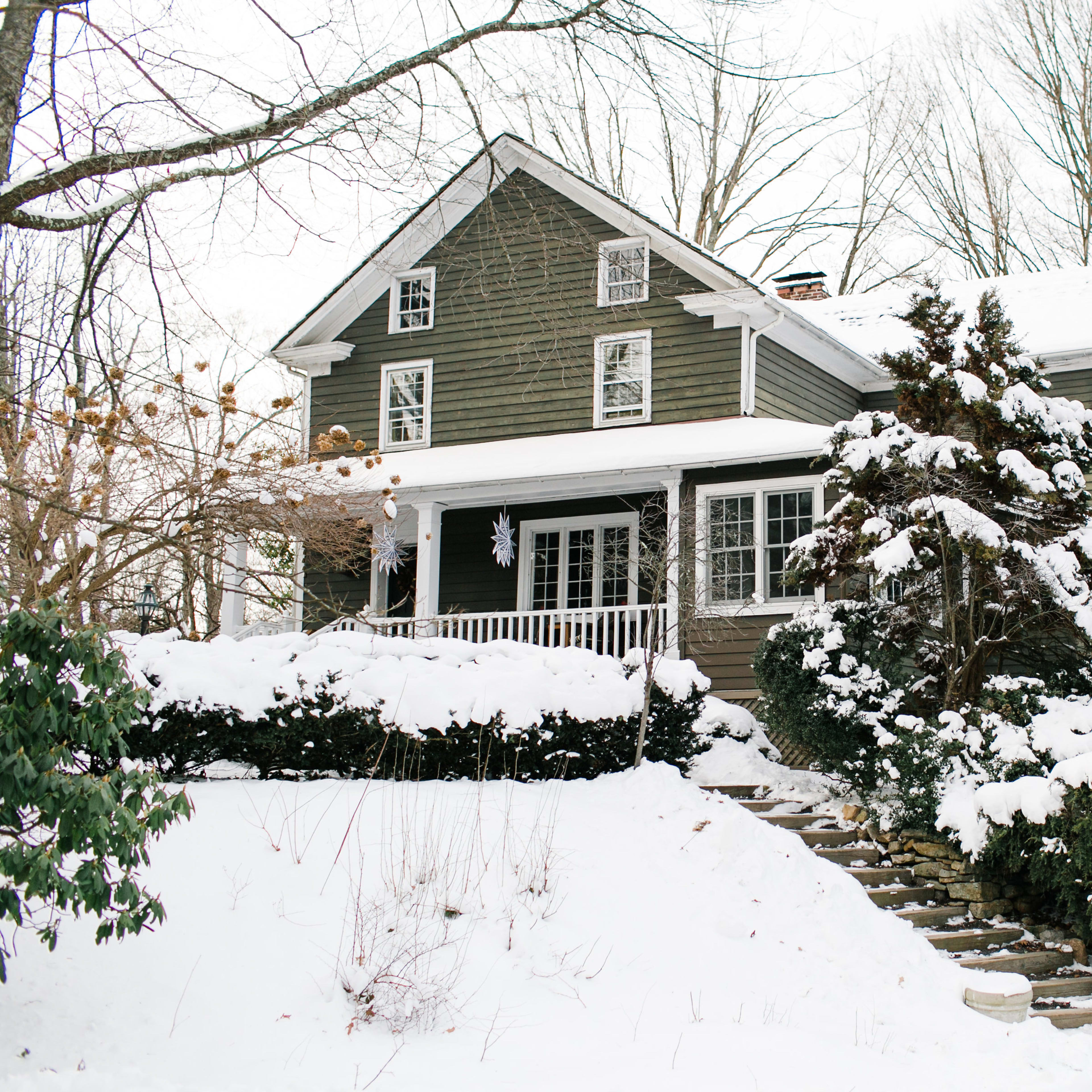 A large, two-story house with a green exterior and a front porch is surrounded by snow-covered trees and shrubs.