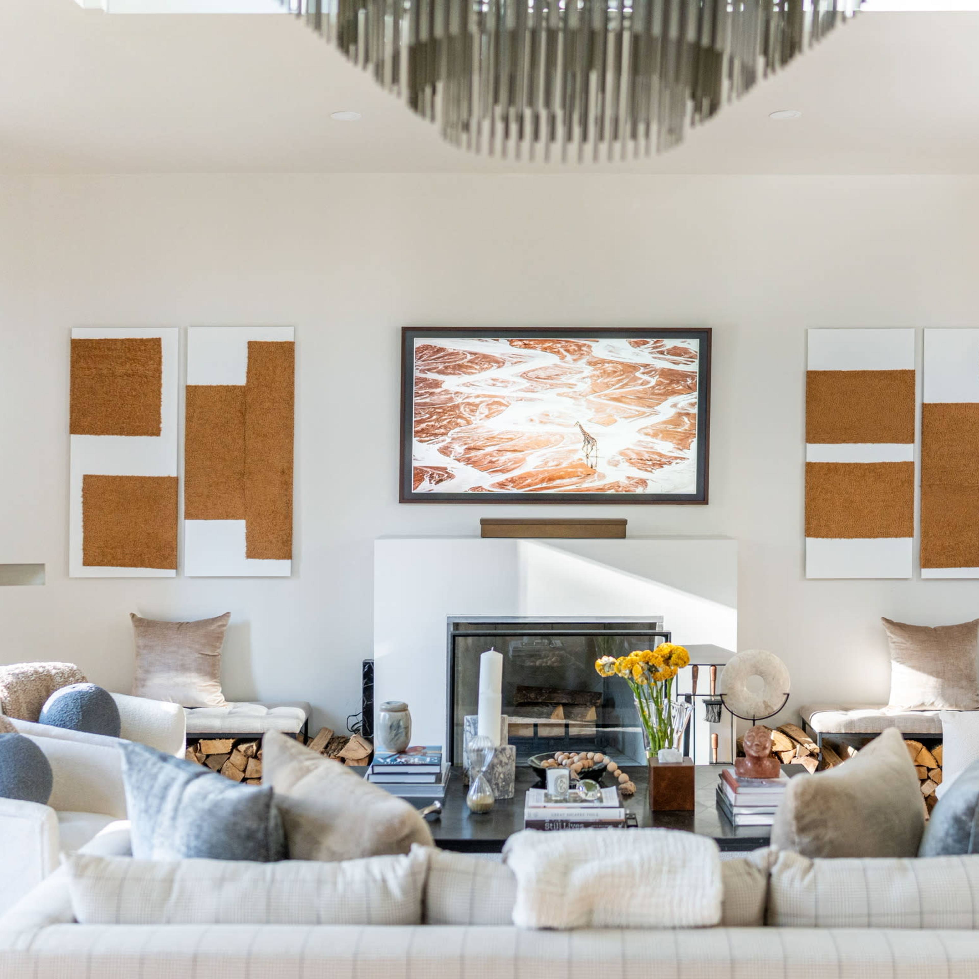 A modern living room with a large television above a fireplace, surrounded by artwork and cozy seating arrangements featuring various cushions and a coffee table.