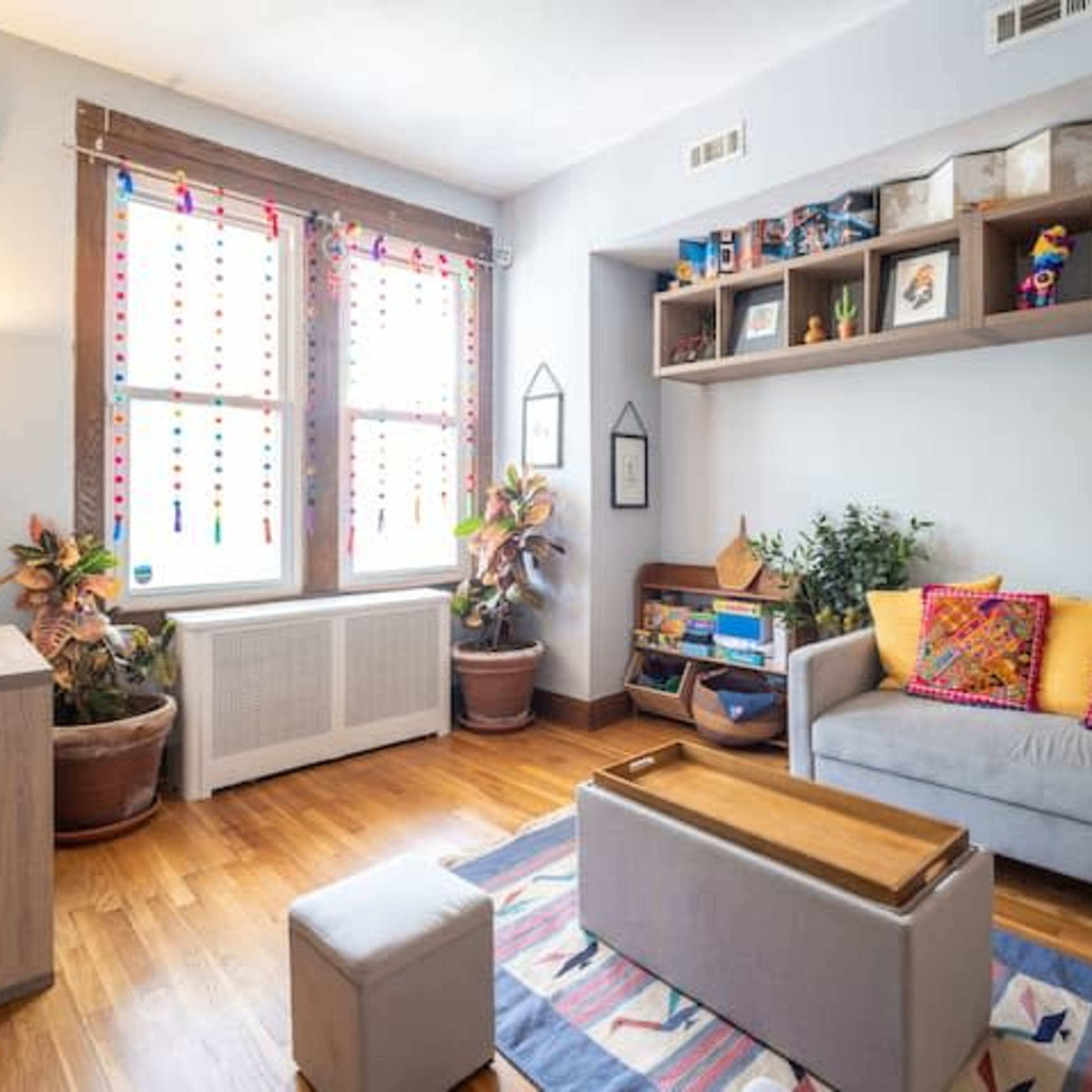 A bright living room featuring a gray sofa adorned with colorful pillows, a wooden coffee table, and shelves displaying various items, with large windows allowing natural light to enter.