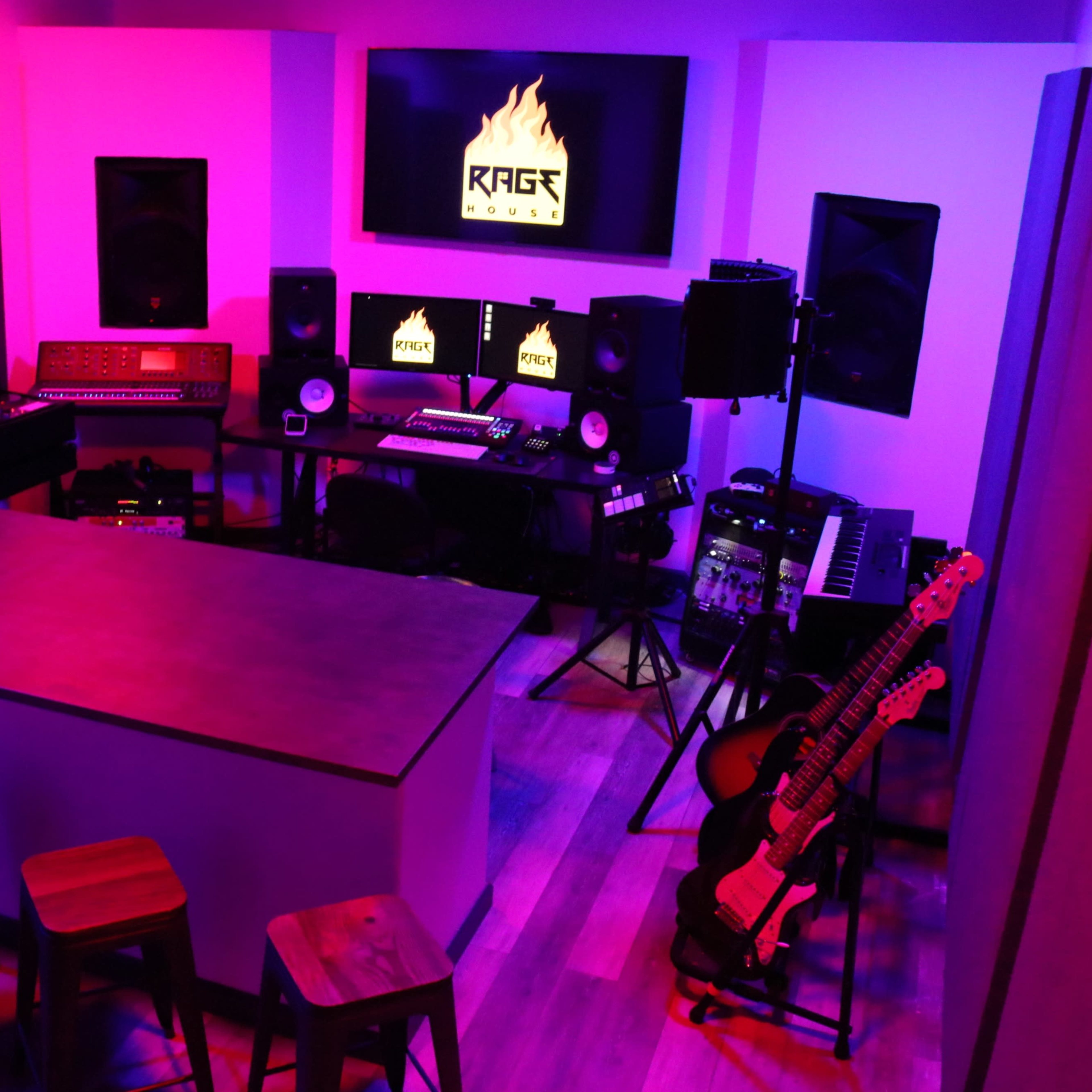 A music production studio featuring a large central table, several audio equipment pieces, and soundproofing panels on the walls, illuminated by purple and pink lighting.