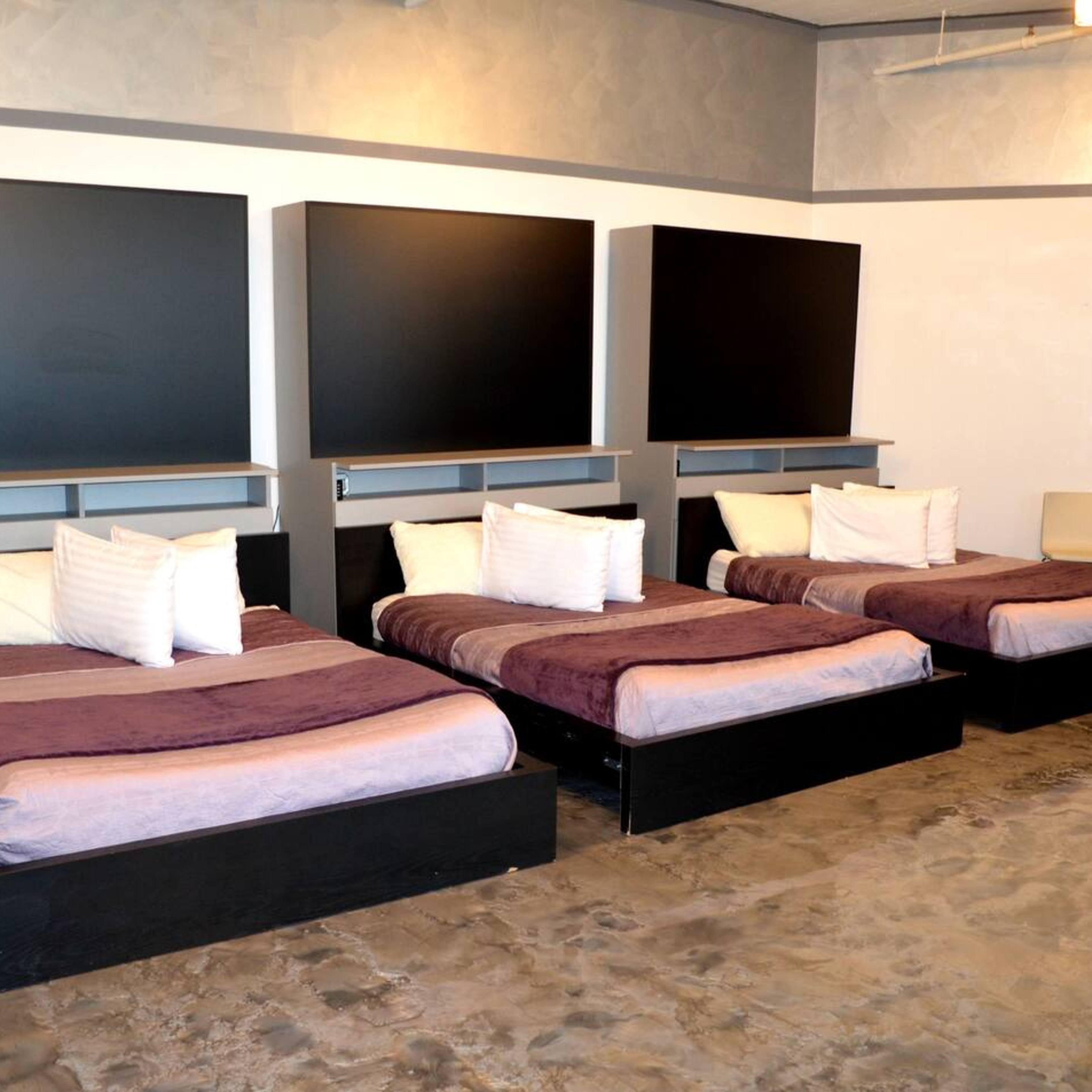 A simple room containing three twin beds with dark wood frames, each made with light bedding and positioned parallel to a wall with two large black panels above them.