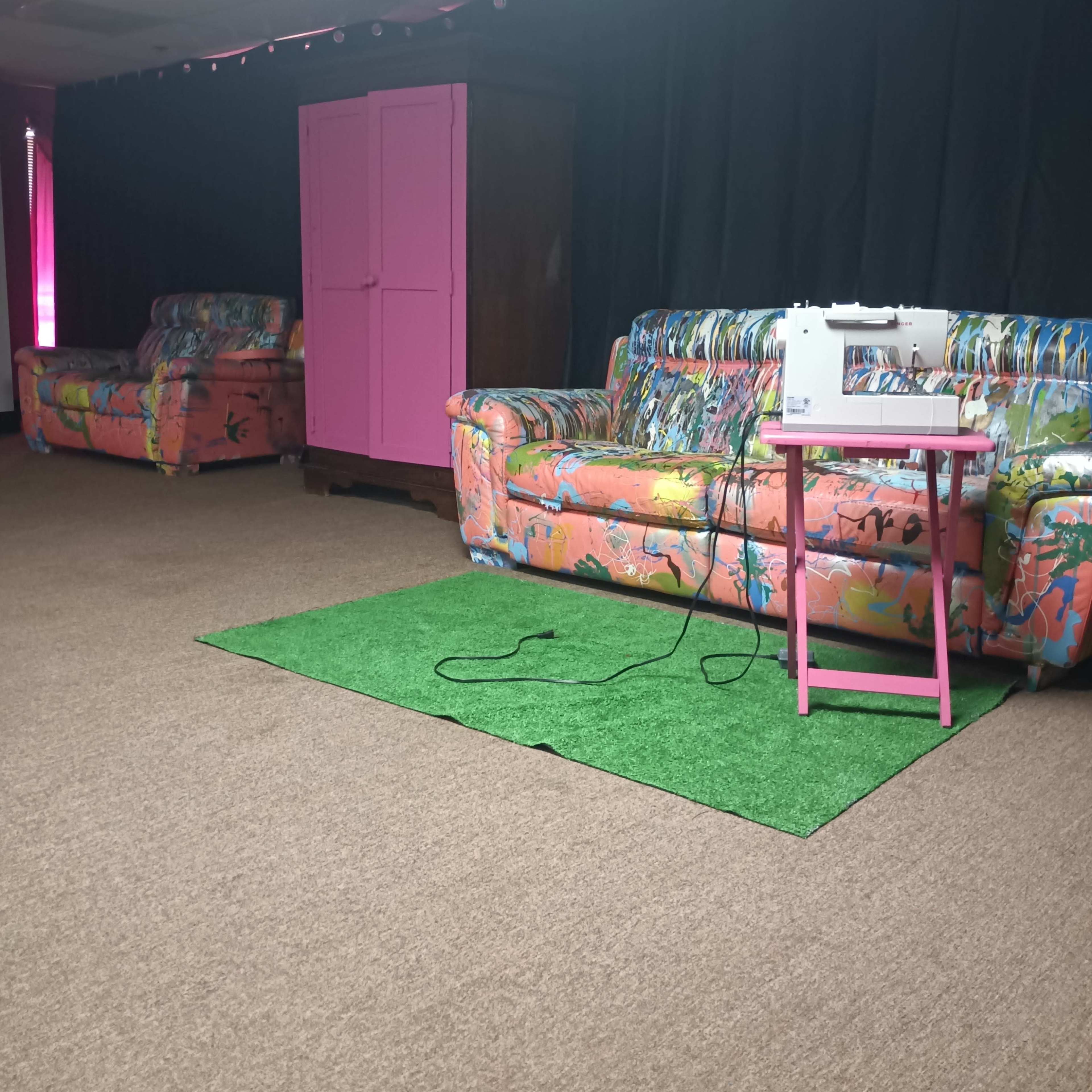 A room with two colorful, patterned sofas, a pink sewing machine on a pink table, and a wooden cabinet against a dark curtain backdrop.