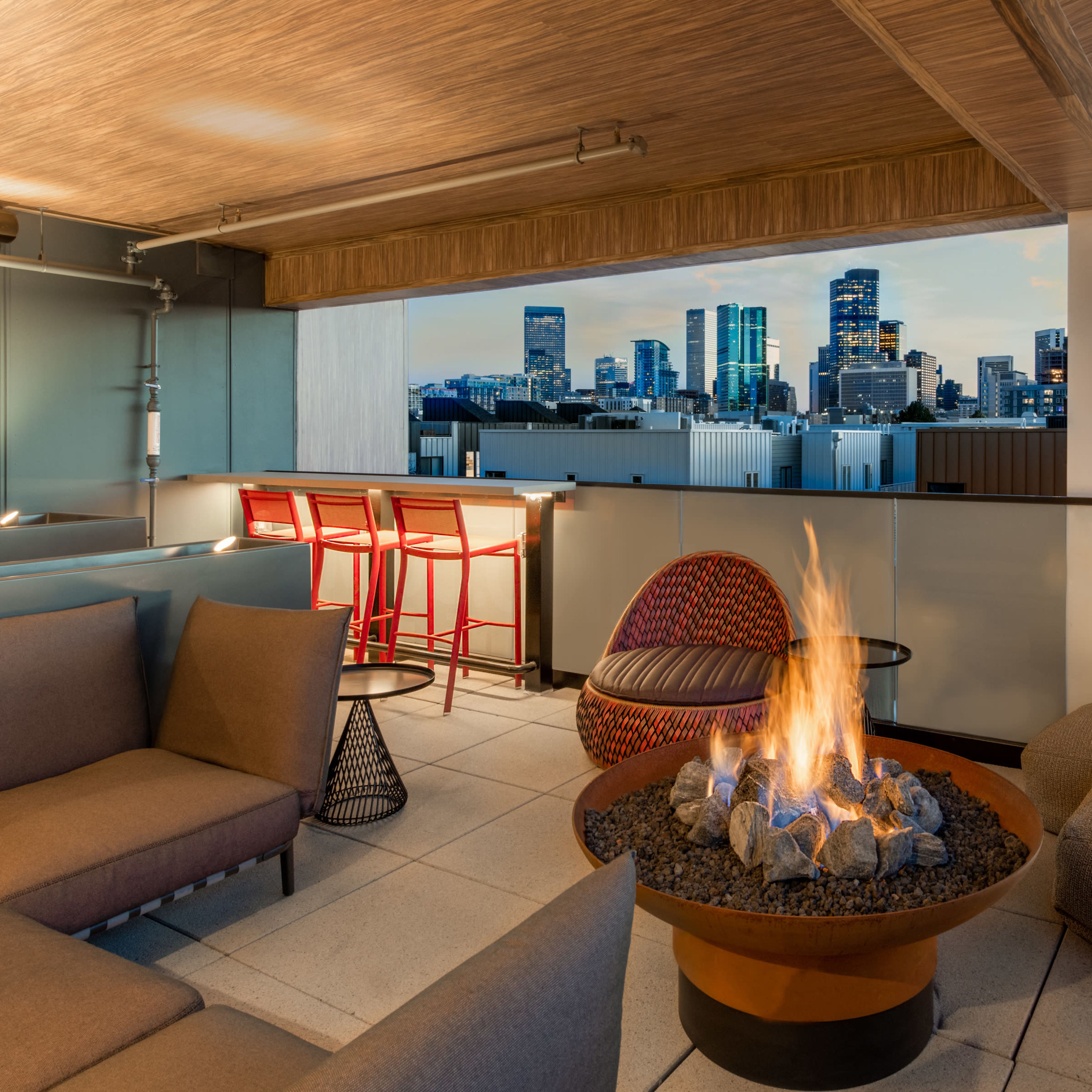 A modern rooftop lounge features a circular fire pit surrounded by comfortable seating, with a skyline view of tall buildings in the background at dusk.