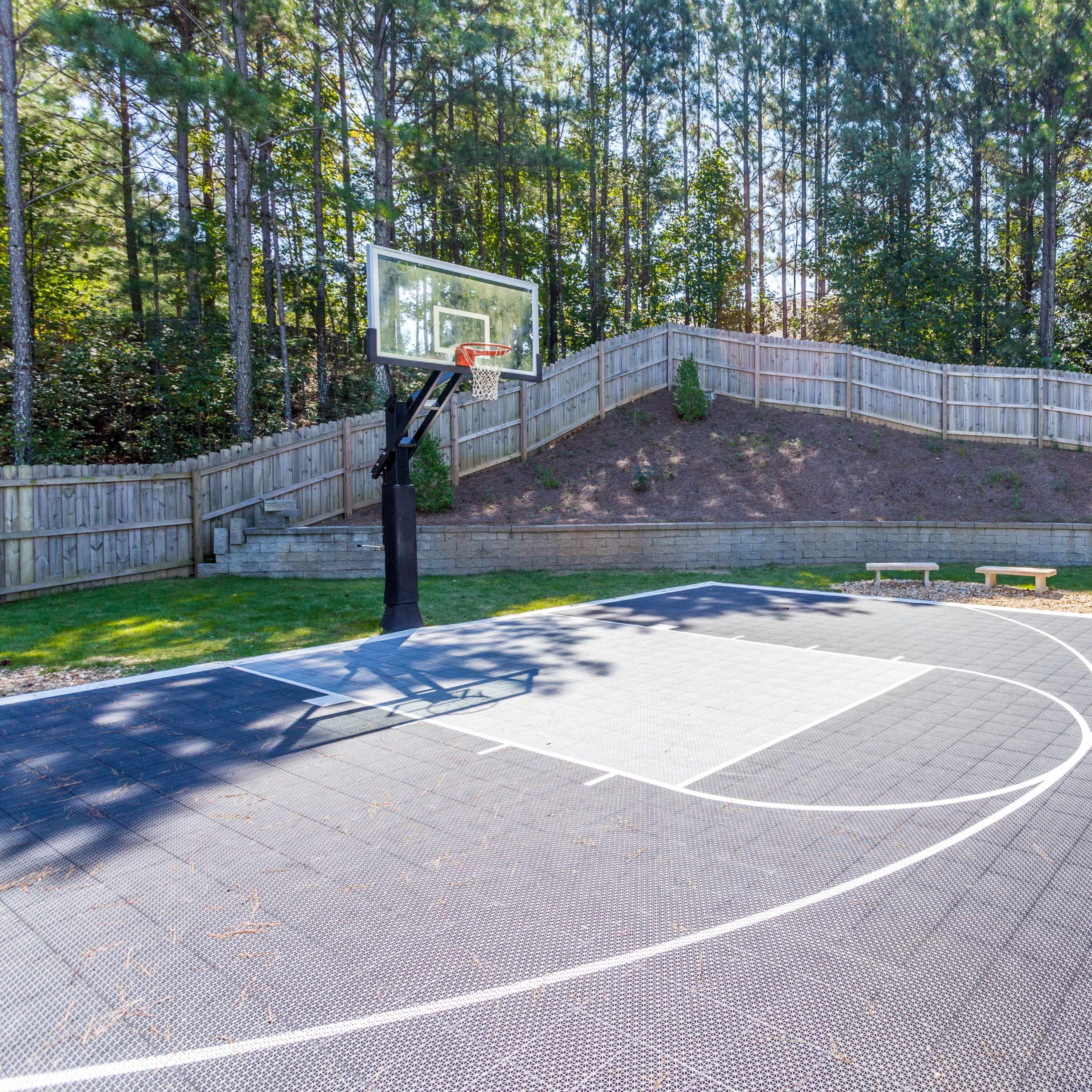 A backyard basketball court with a hoop, surrounded by trees and a wooden fence.