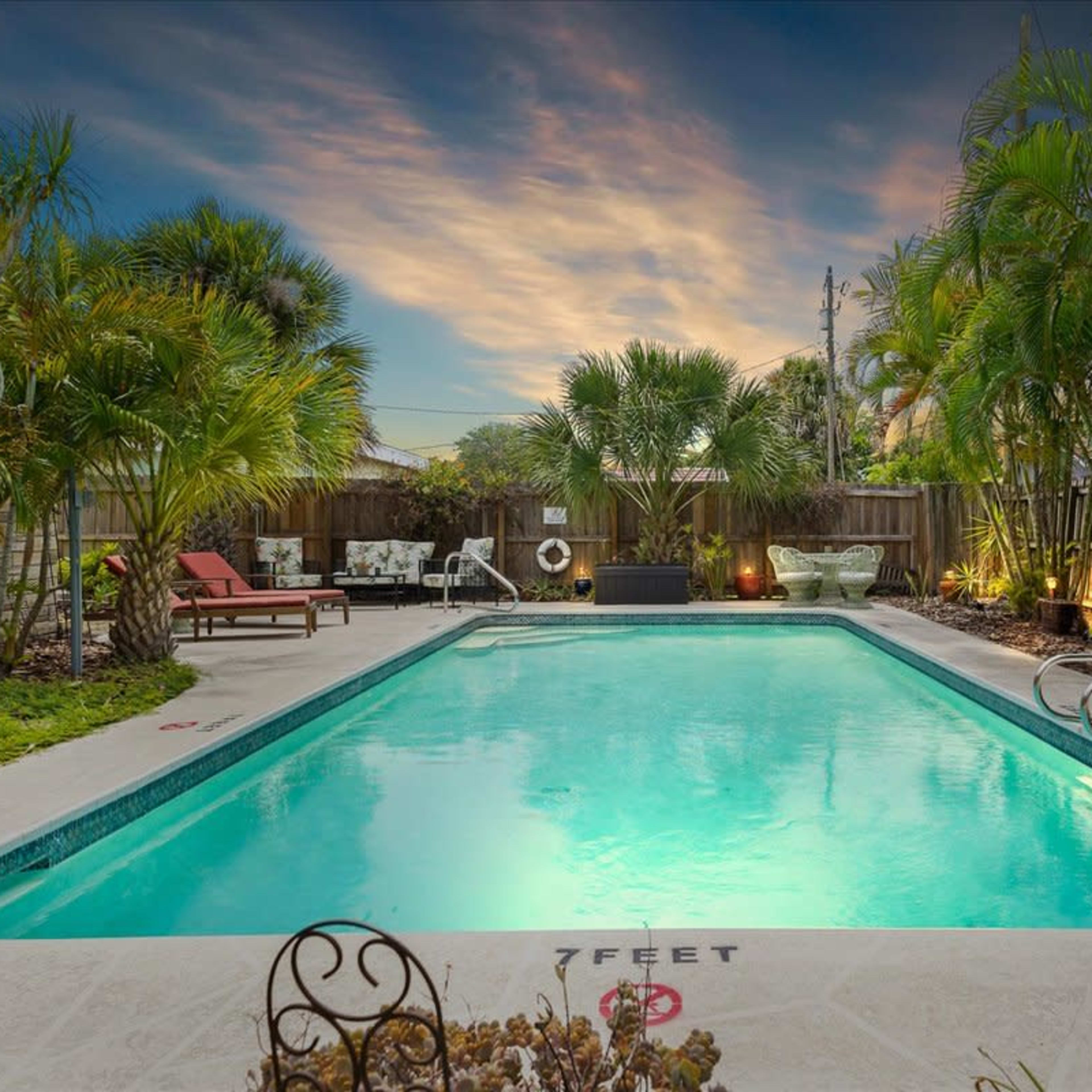 A rectangular swimming pool surrounded by palm trees and lounge chairs under a colorful sunset sky.