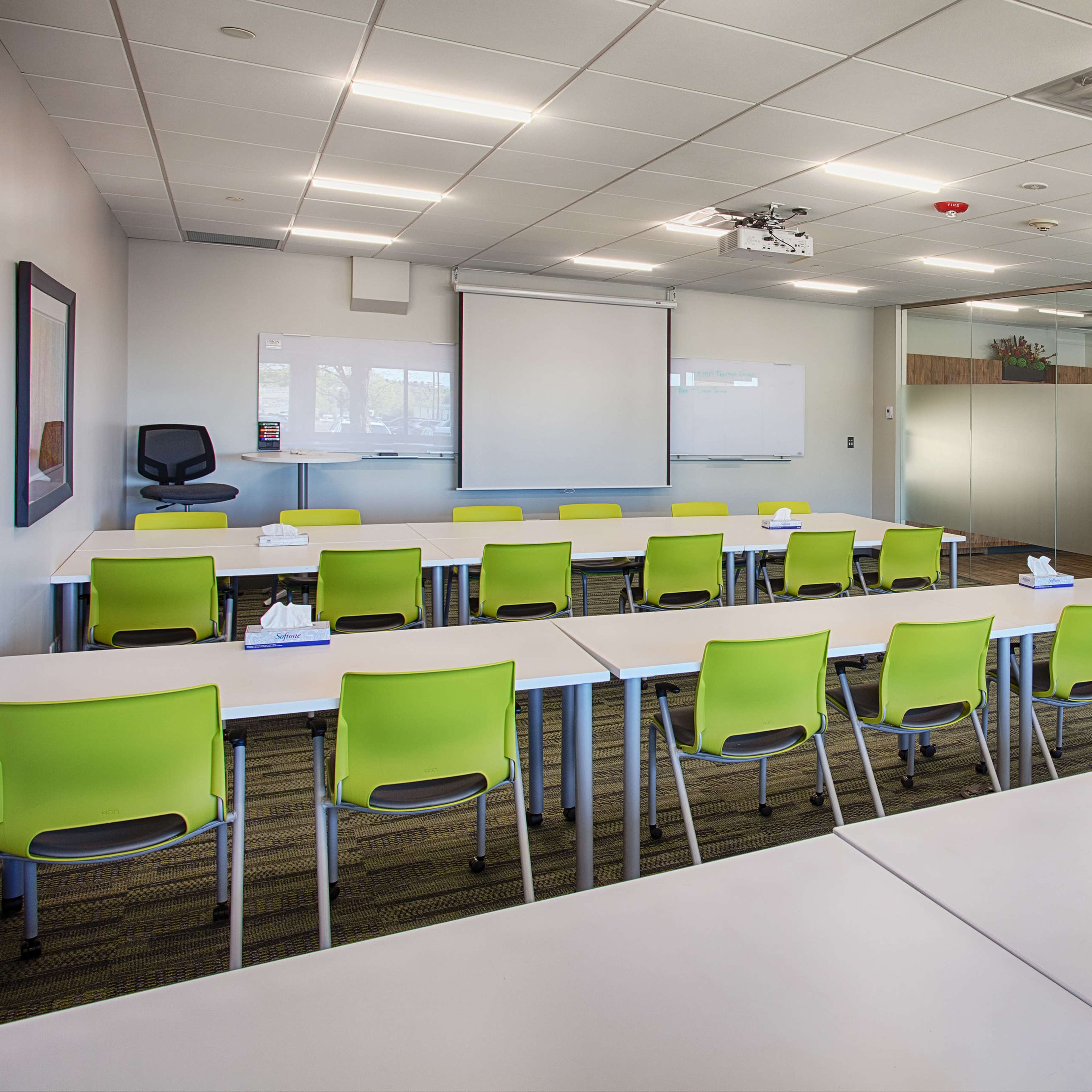 A classroom features several white tables arranged in rows with green chairs and a whiteboard at the front.