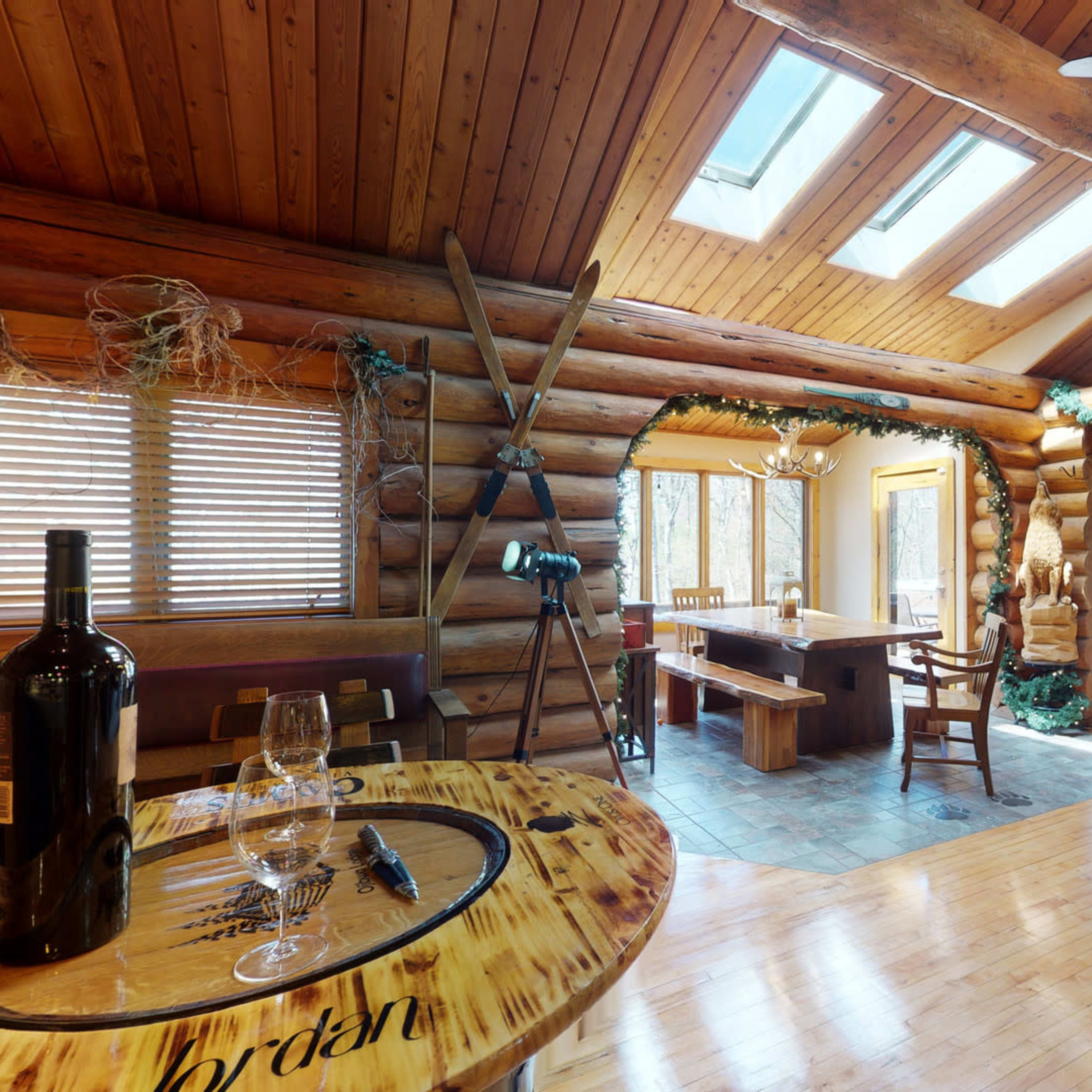 A spacious log cabin interior features a dining area with a wooden table and chairs, large windows, and a living space with musical instruments and holiday decorations.